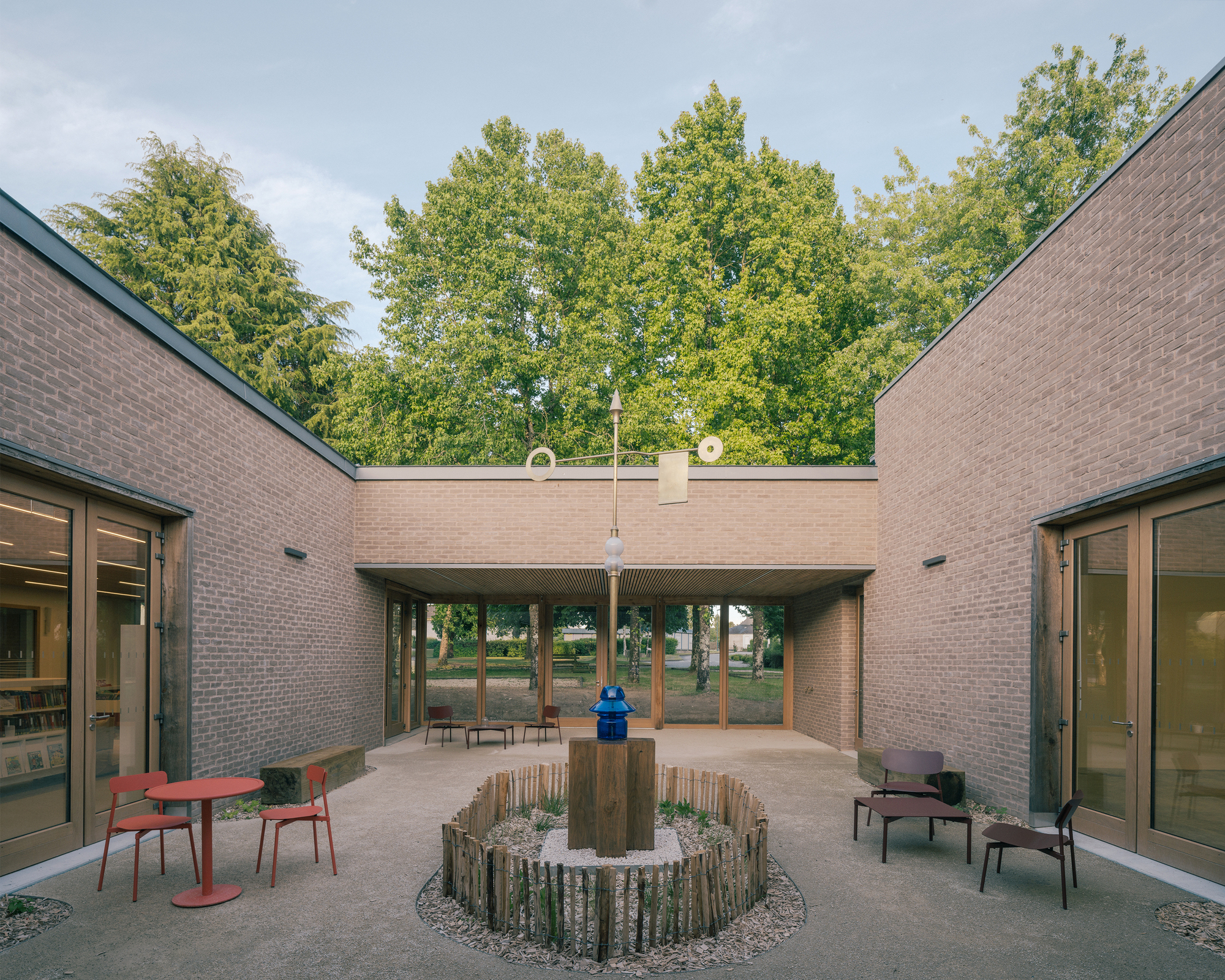 Brick courtyard with a central planted ring of timber stakes, a blue lantern on a post, and red chairs under tall trees