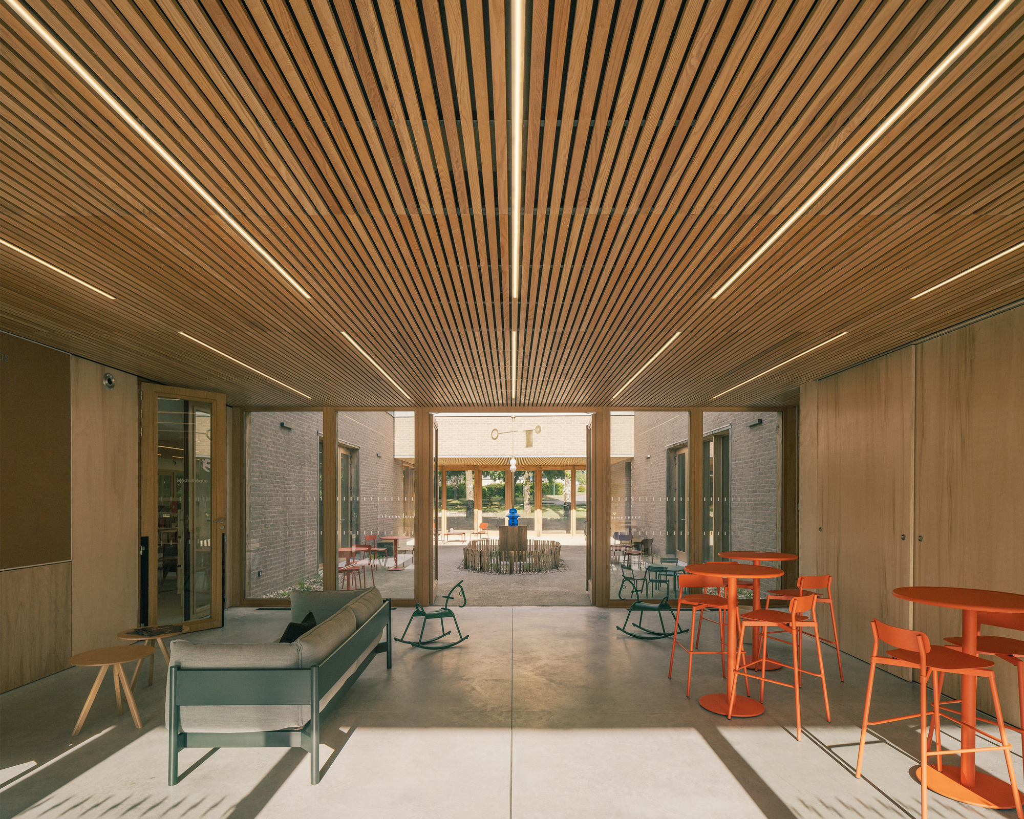 Covered gathering space with a timber slat ceiling and linear lights, orange stools and a sofa, opening toward the courtyard in afternoon sun
