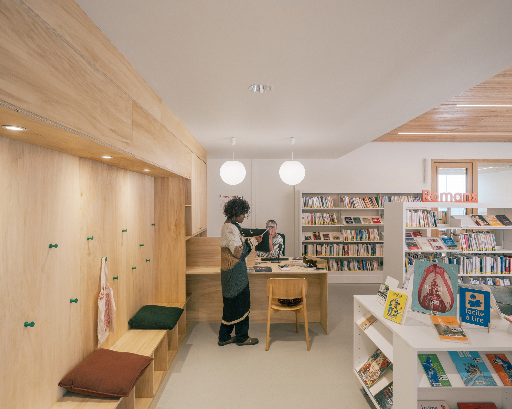 Library interior with a plywood wall of pegs and bench seating, a librarian at the desk, and a visitor standing beside shelves