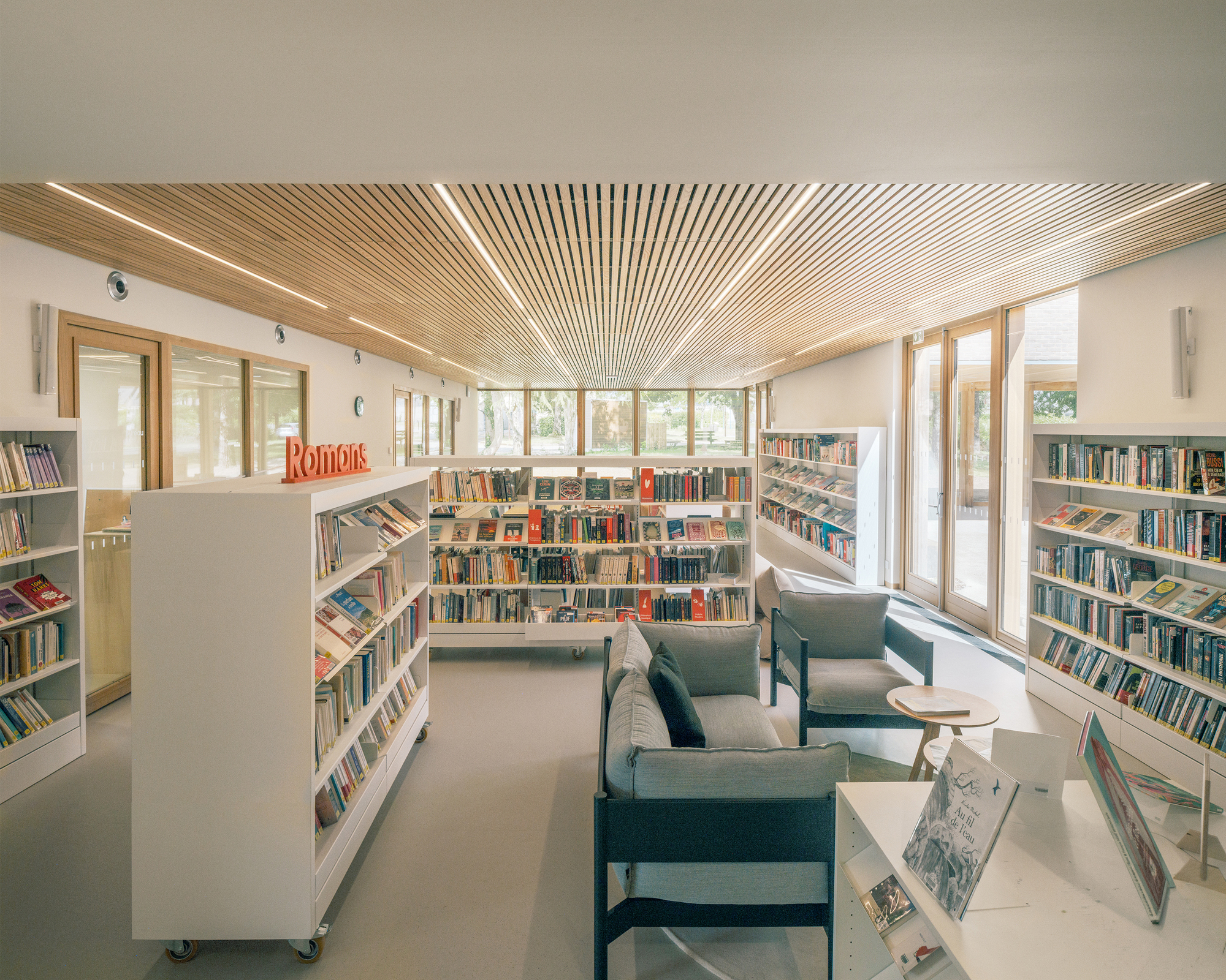 Reading area with grey armchairs and mobile white shelving beneath a slatted timber ceiling, daylight entering through tall glazed doors
