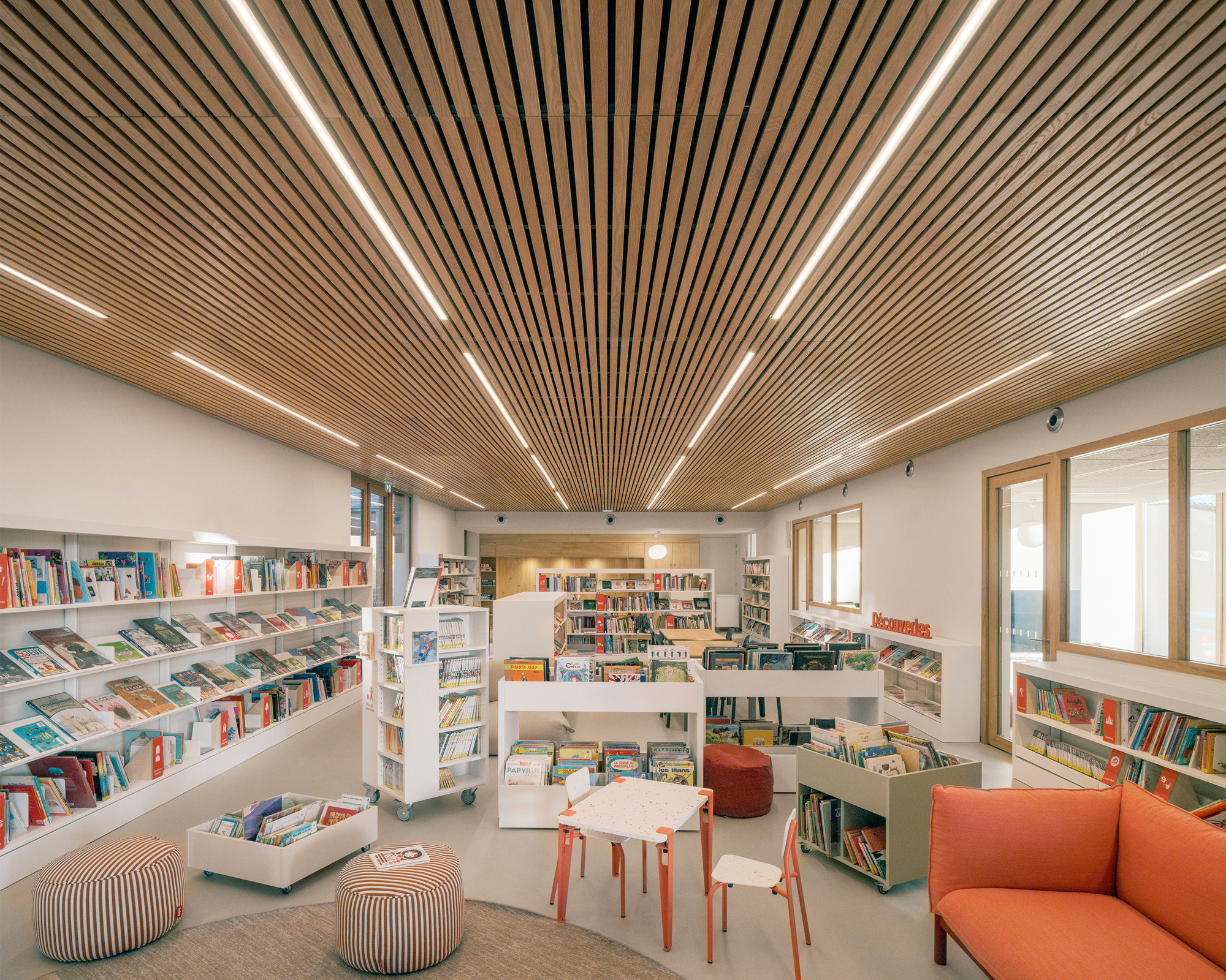 Children's library zone with orange sofa, striped poufs and a small table under a slatted timber ceiling with linear lights