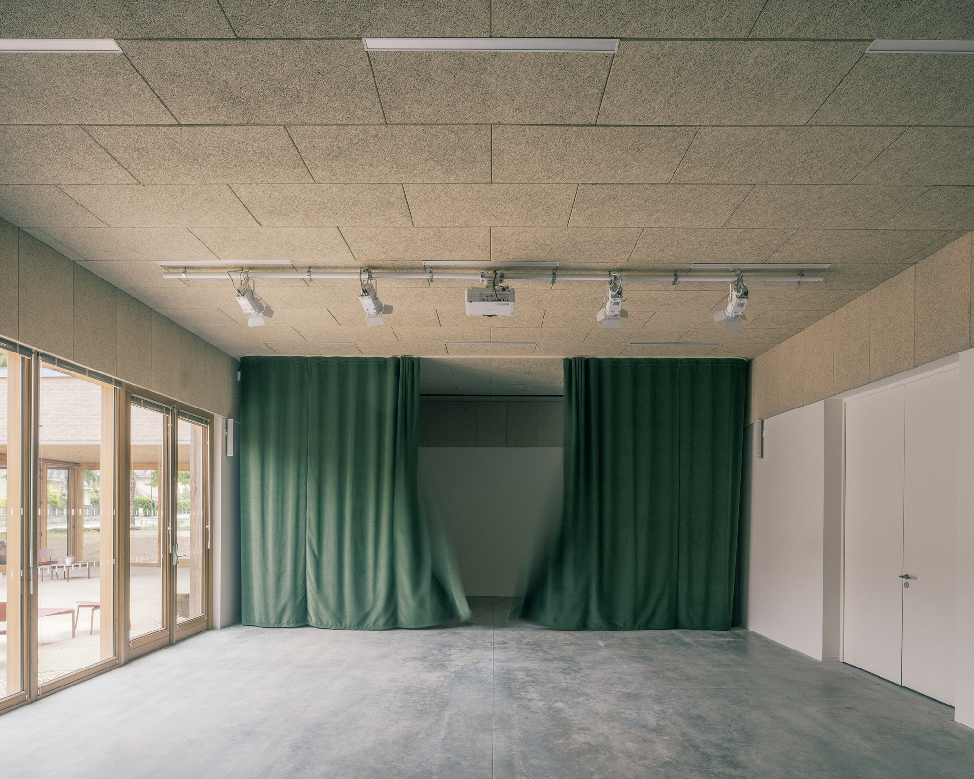 Multipurpose room with green velvet curtains half drawn, ceiling projector on a track and bare concrete floor beside timber-framed glazing