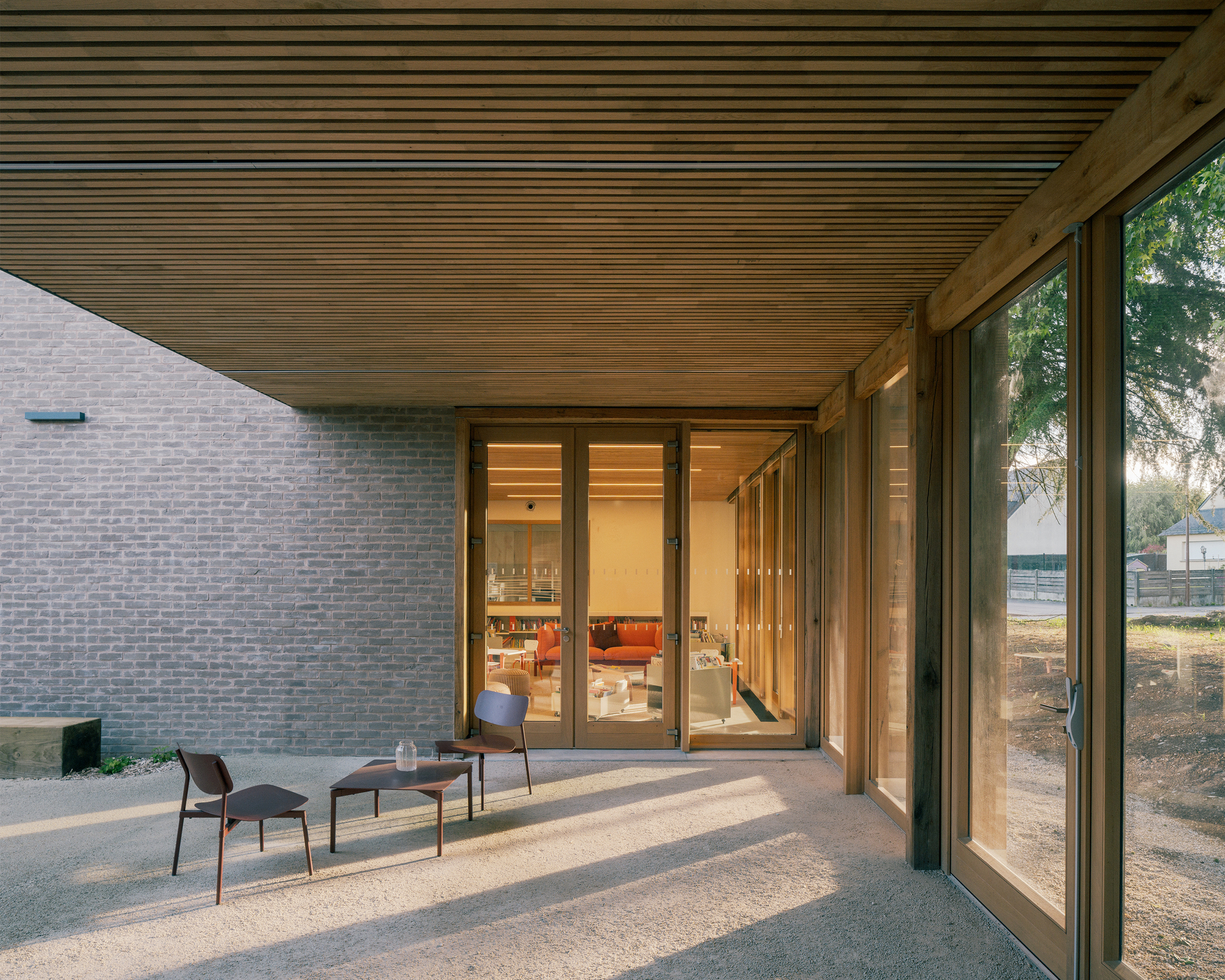 Covered gravel terrace with two small chairs and a table, sunlight cutting under a timber slat soffit beside a grey brick wall