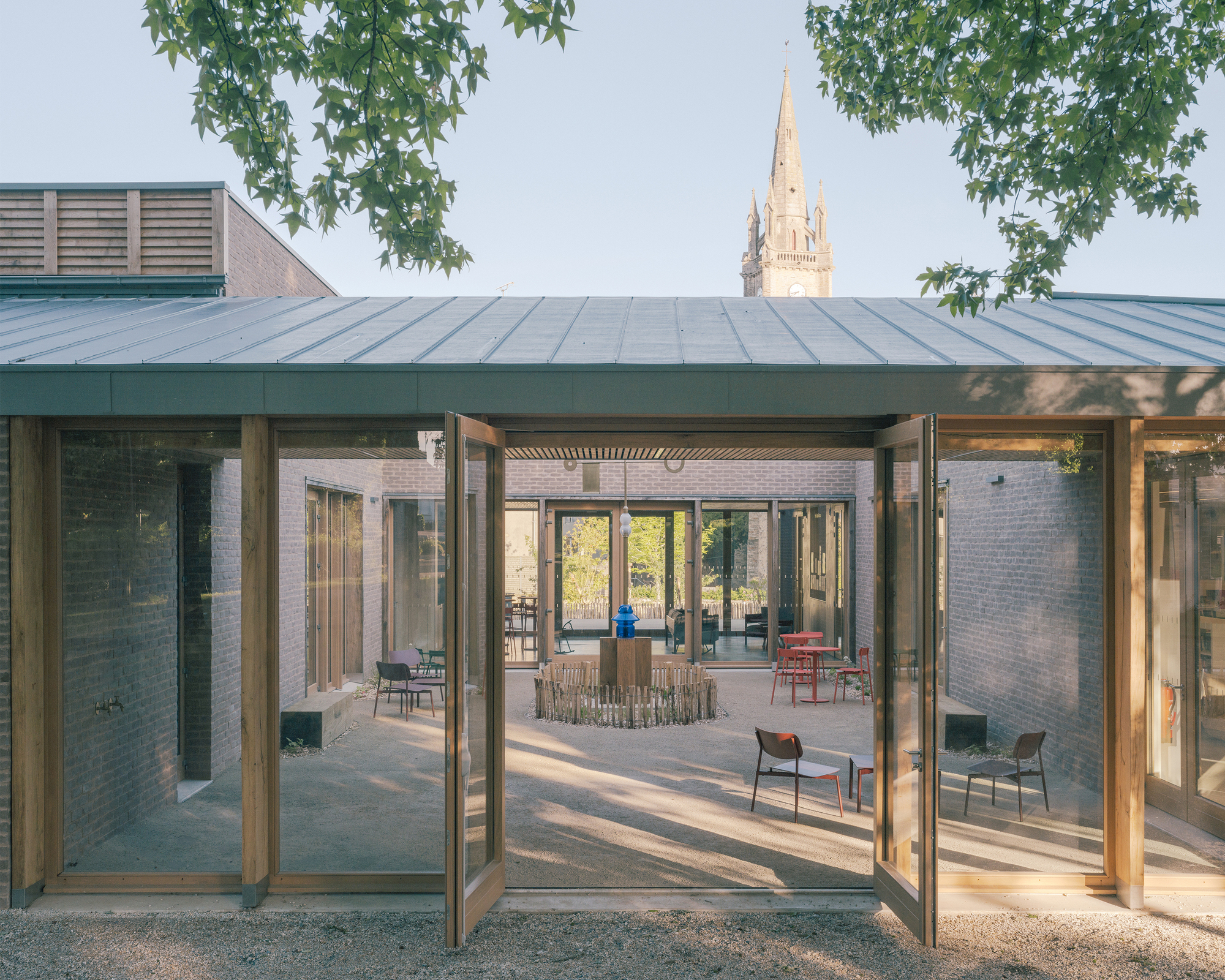 Courtyard seen through open glazed doors beneath a zinc roof, with a church spire rising behind leafy branches in clear sky
