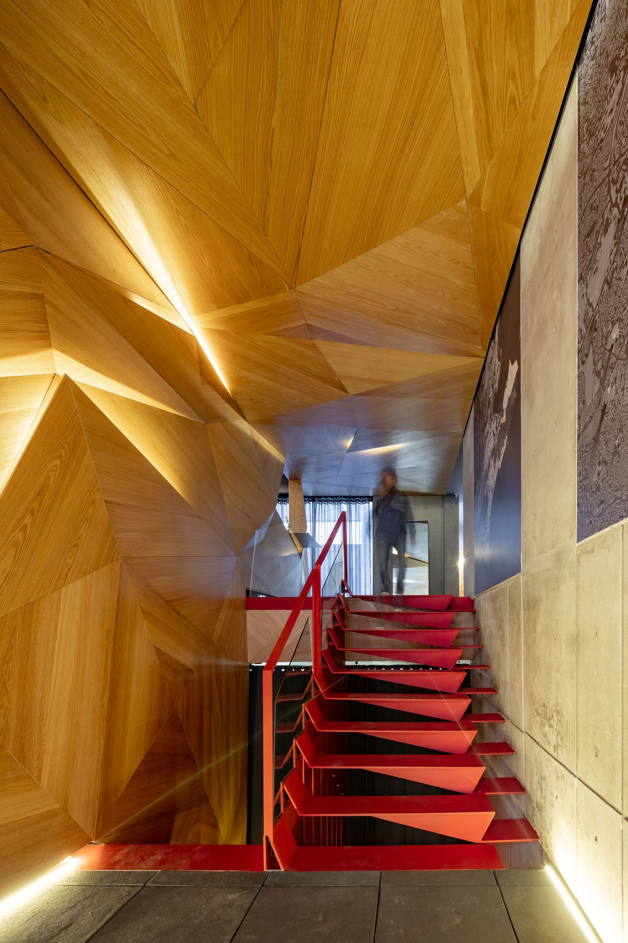 Red steel stair descending beside a faceted oak-veneer ceiling and wall, with a blurred figure standing at the landing