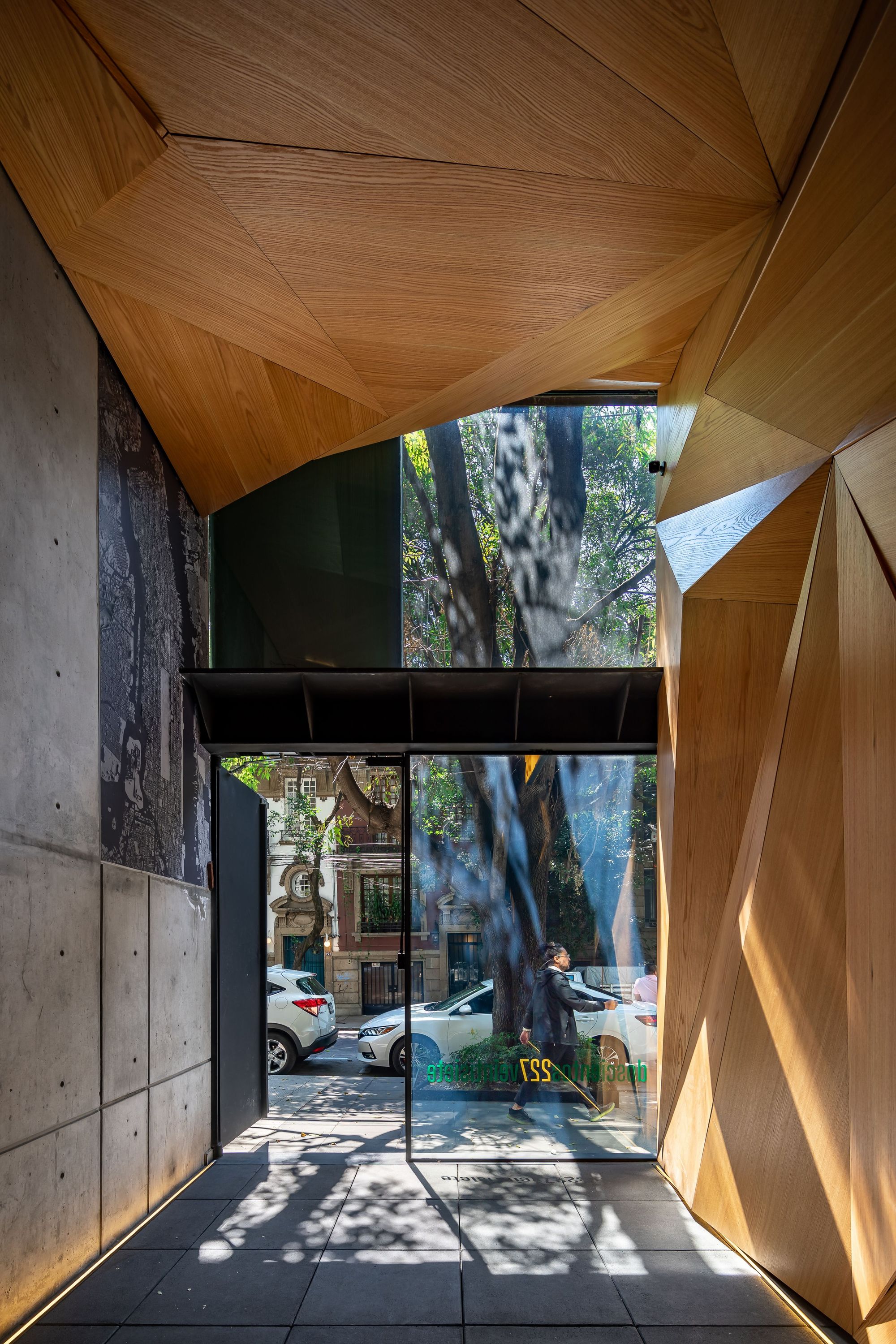 Entry passage with faceted timber ceiling, board-marked concrete wall and glass front framing a street tree and a passing pedestrian