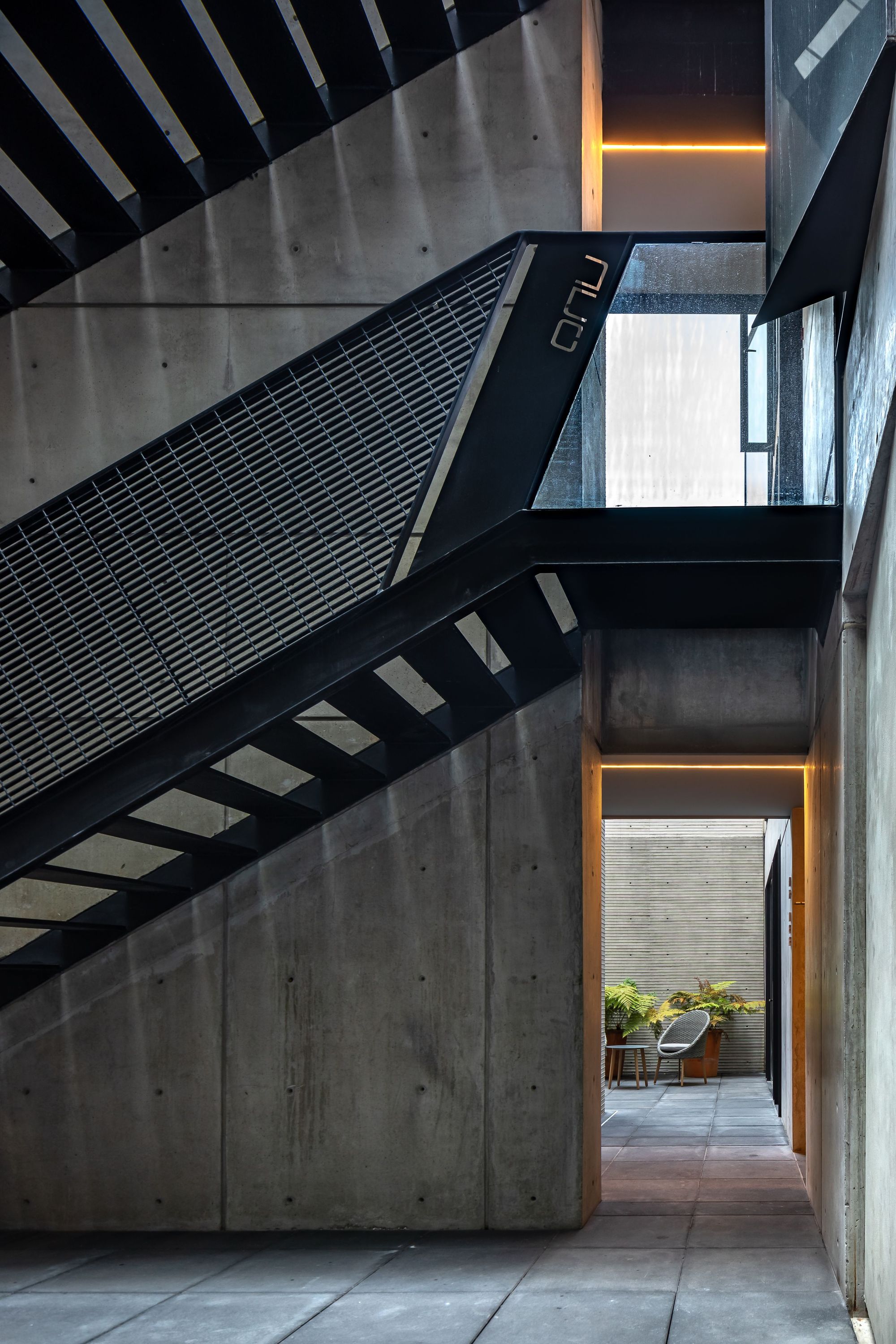 Concrete stairwell with black steel stair and mesh guard, opening onto a small courtyard with potted ferns and a chair
