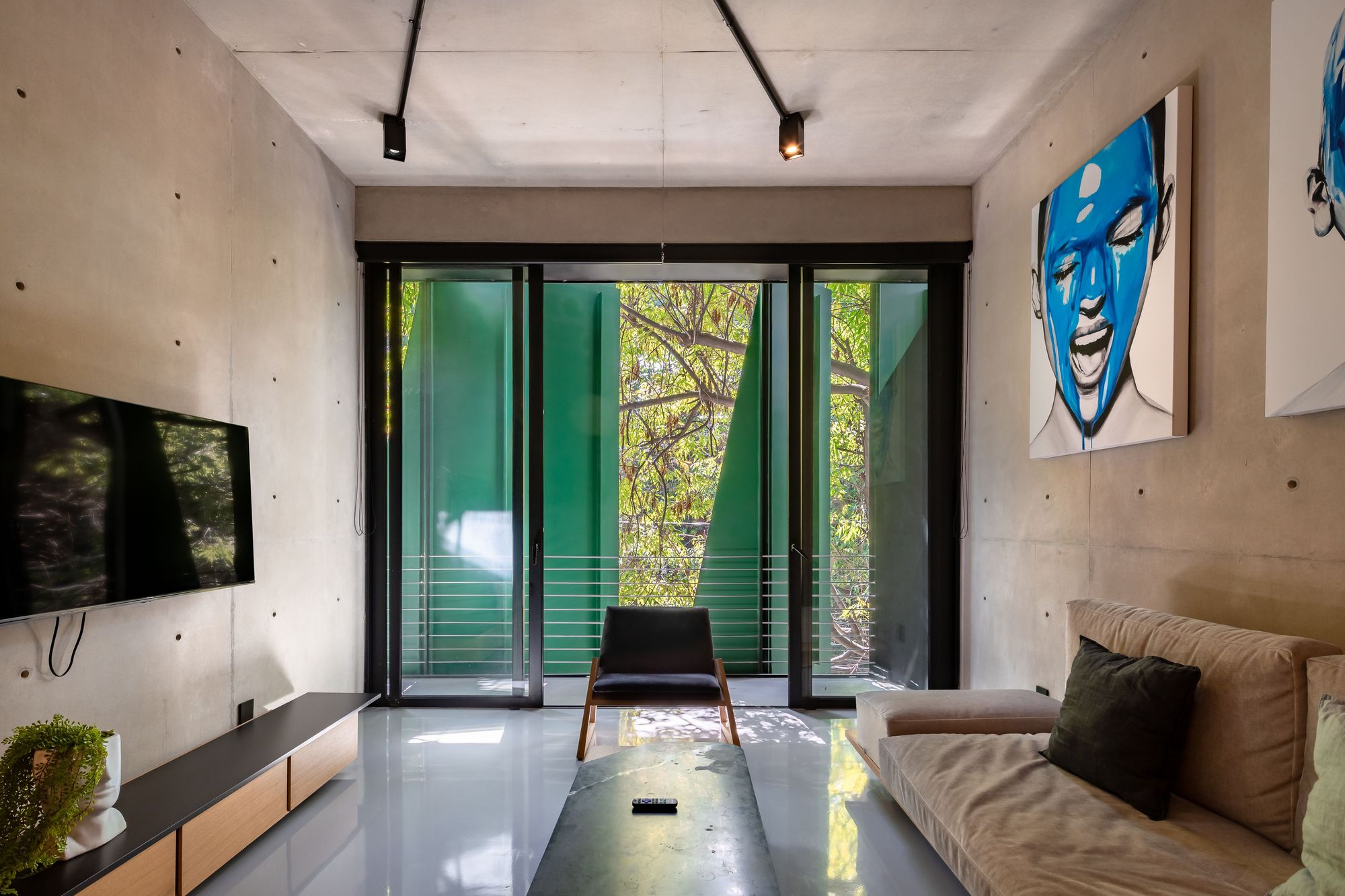 Living room with exposed concrete walls, sofa and blue portrait painting, facing glass doors onto green fins and tree branches