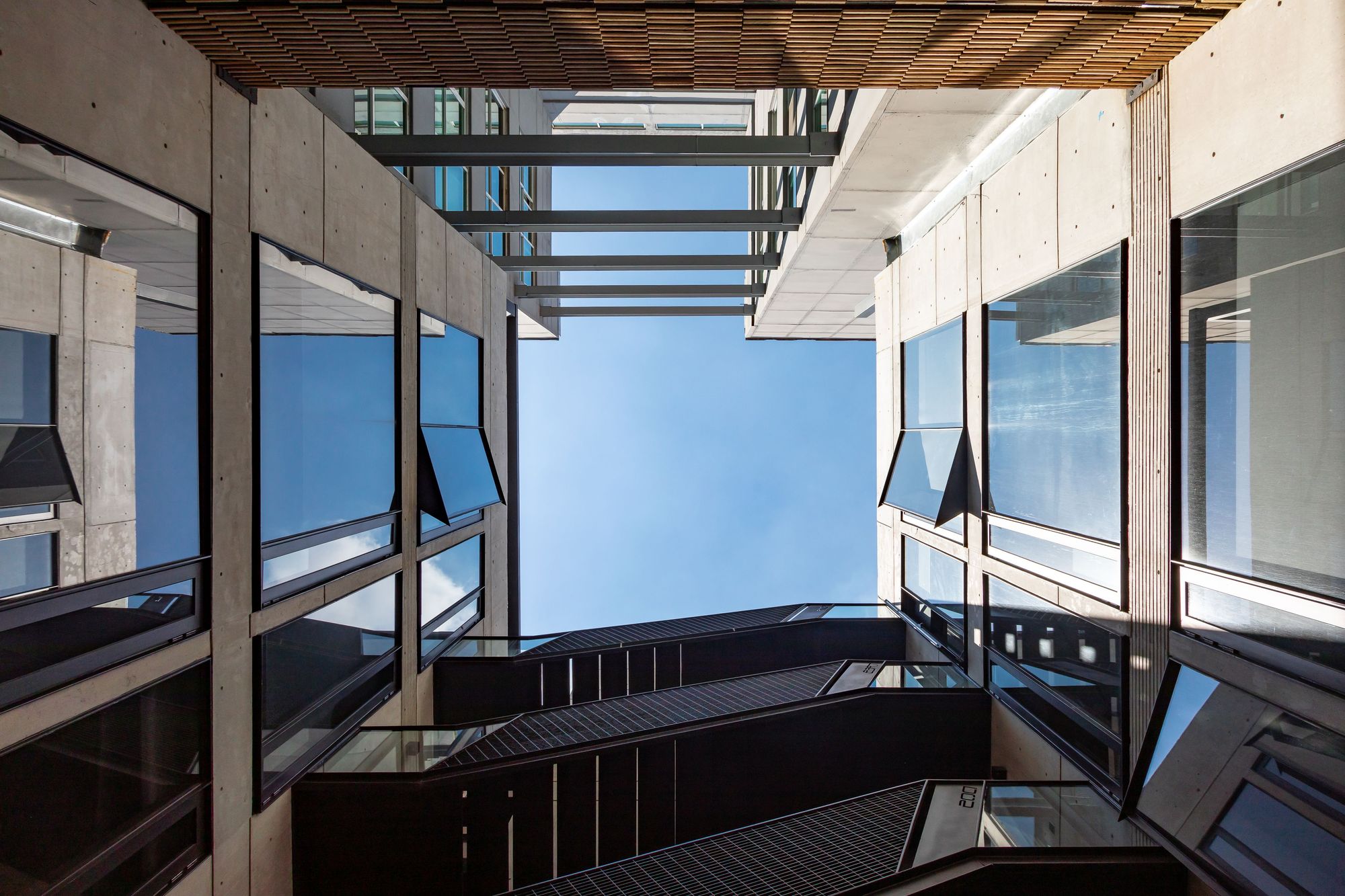Upward view from a narrow light well, with concrete panel walls, tilted glazing and dark steel stair runs against a blue sky
