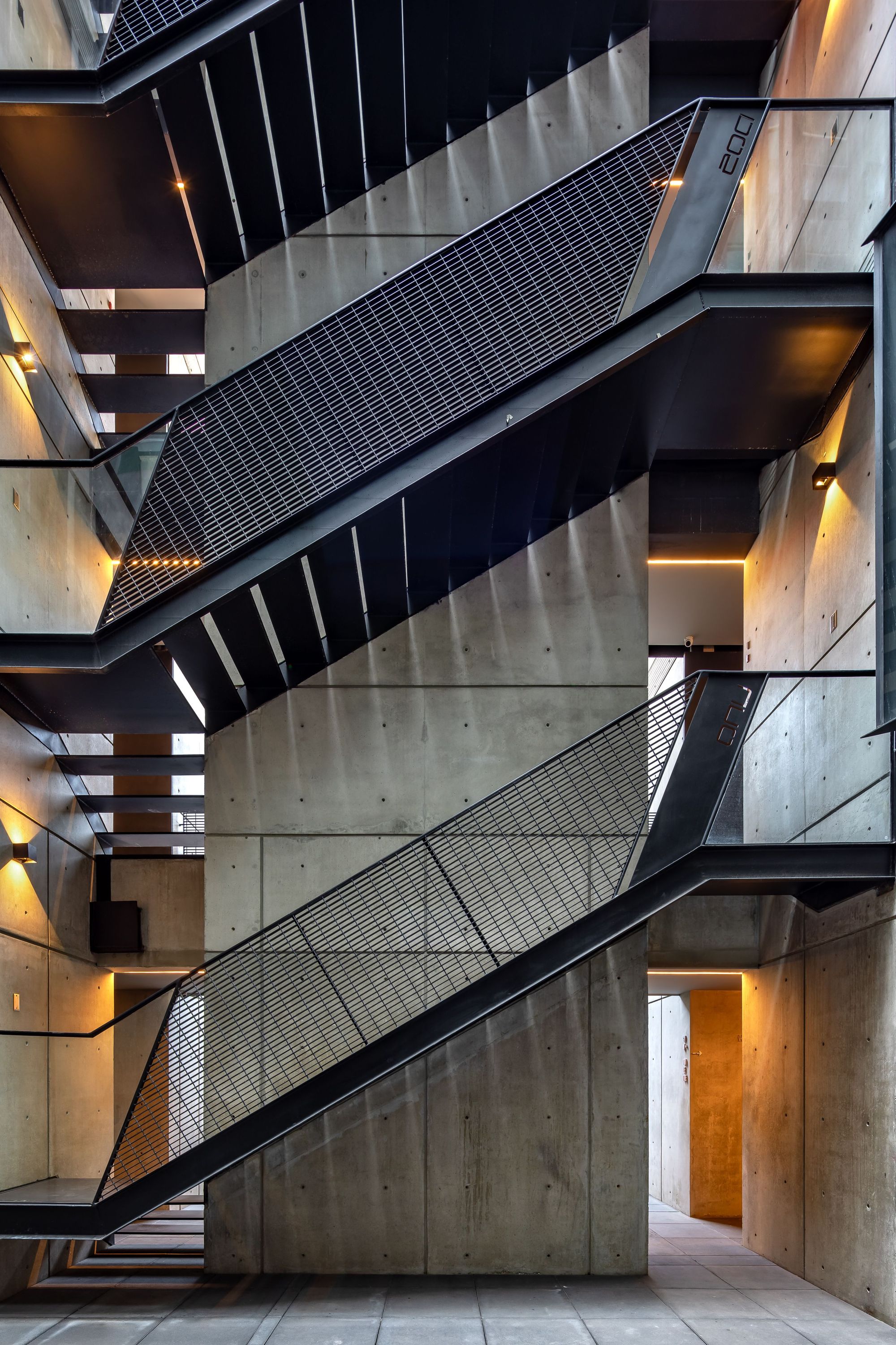 Zigzag steel staircases with mesh balustrades climbing a board-marked concrete wall, lit by warm wall lamps at dusk