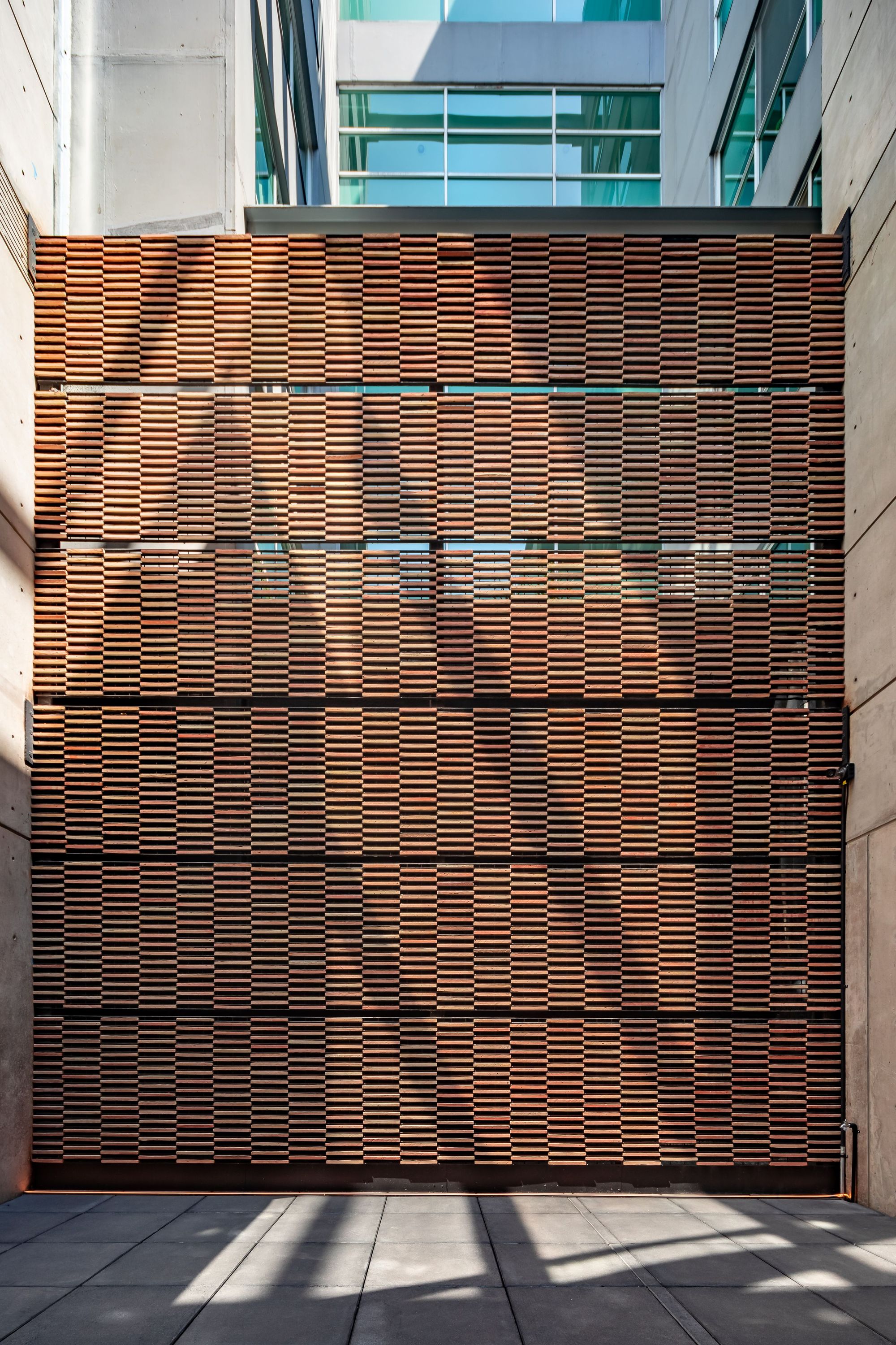 Courtyard wall clad in stacked terracotta slats with shadows of nearby buildings falling across the paving in afternoon sun