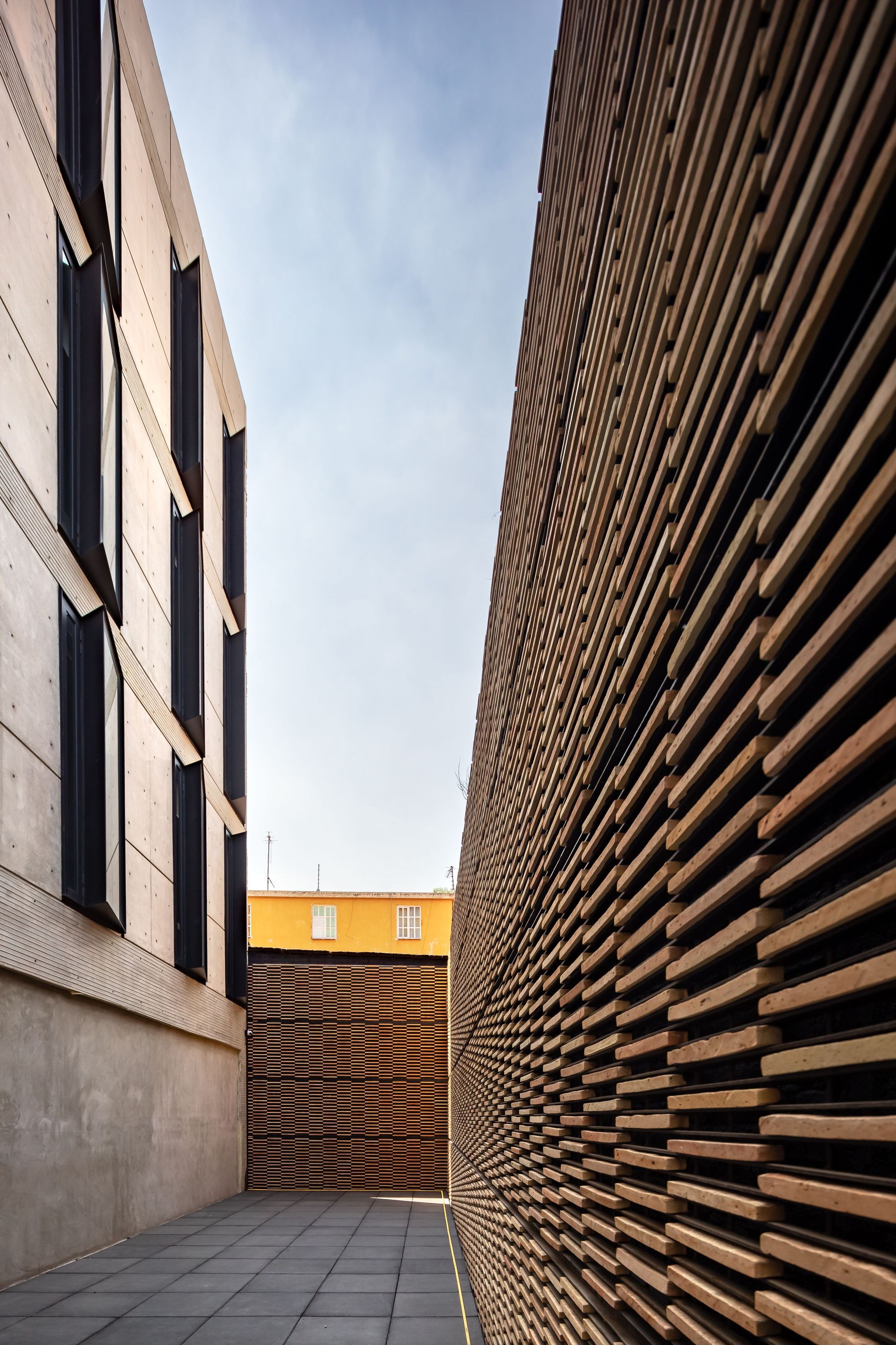 Narrow alley between a board-marked concrete wall with dark window frames and a timber slat screen, under a pale blue sky