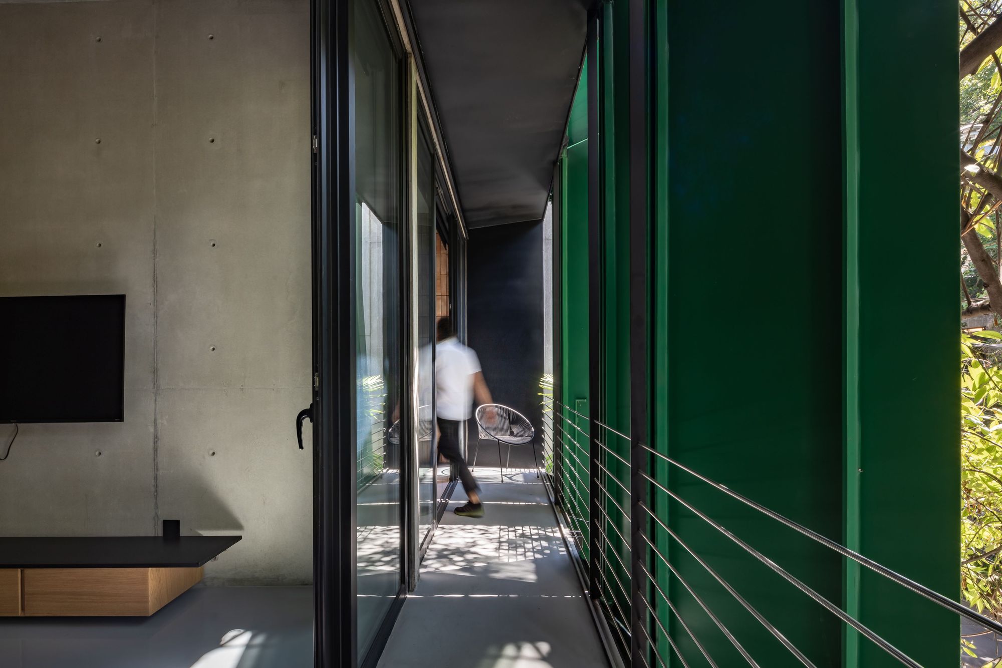 Balcony corridor between a sliding glass wall and a green panel with cable railing, a man walking past in dappled sunlight