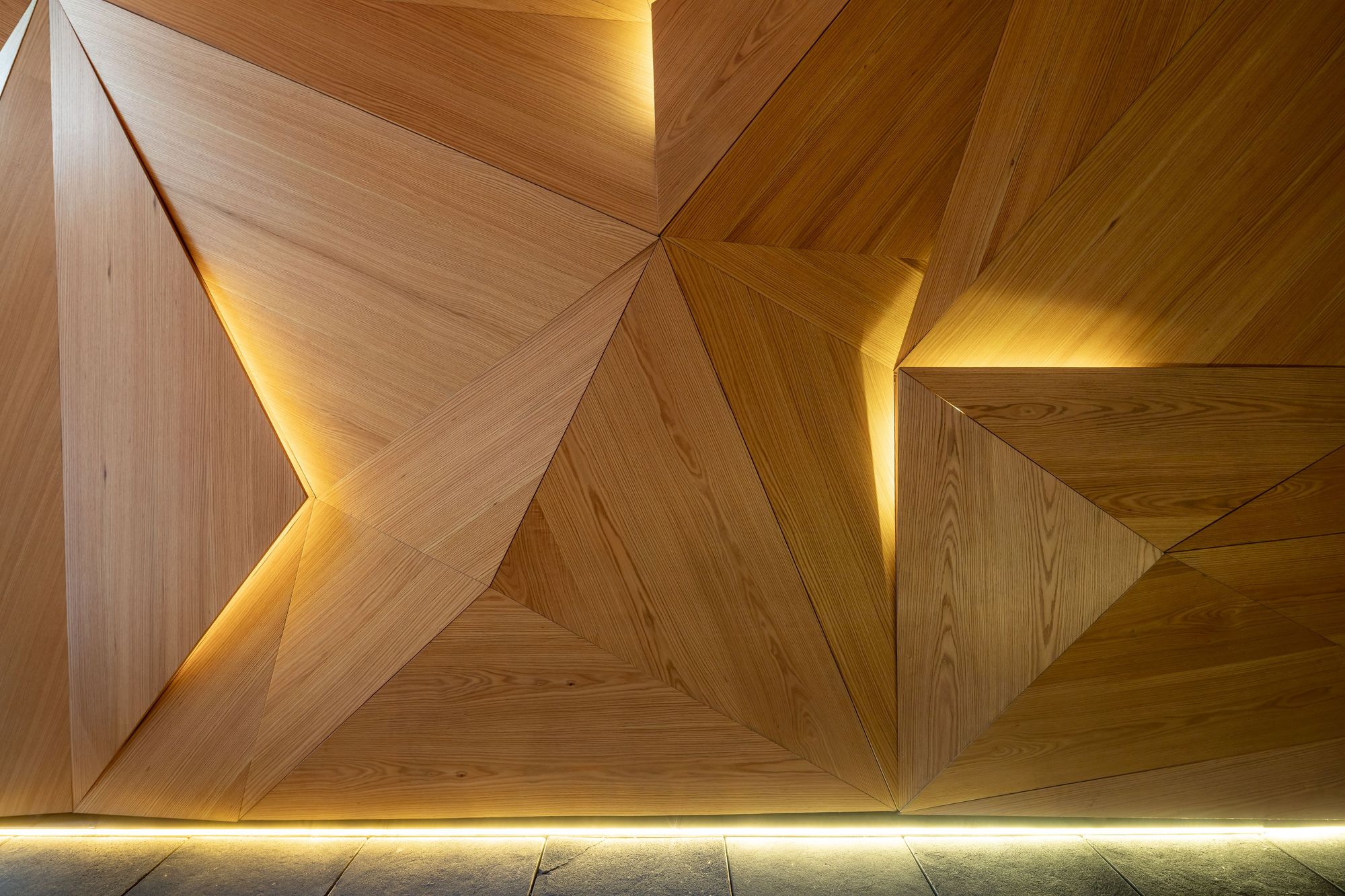 Faceted wall of triangular oak panels with warm concealed light along the seams above a slate floor