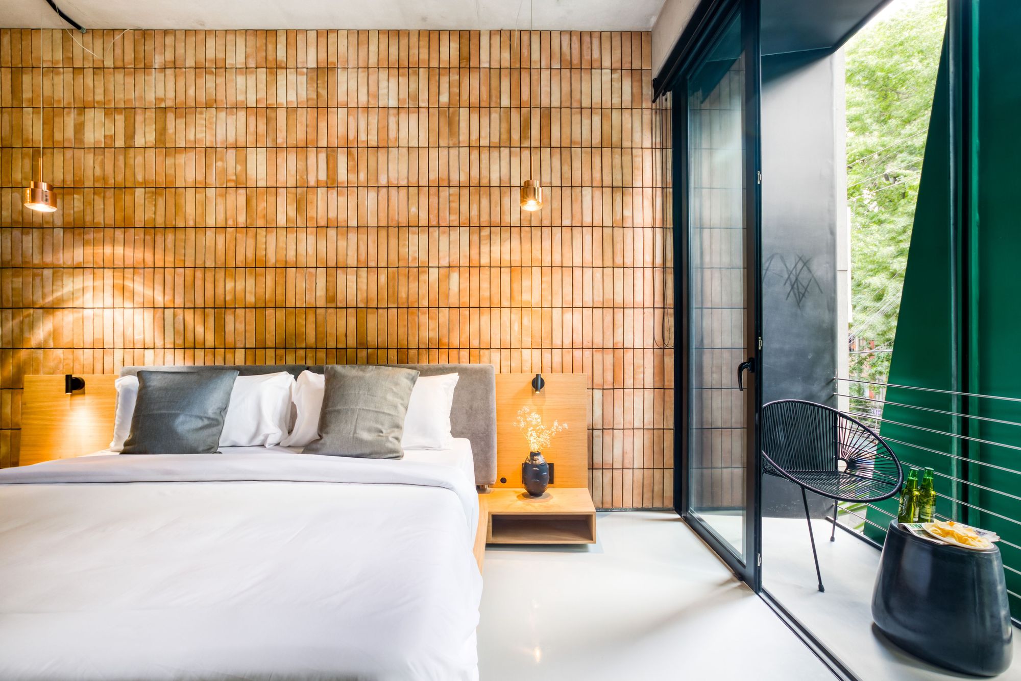 Bedroom with a brick-slip headboard wall, copper pendants and a glass door opening to a balcony chair among green leaves