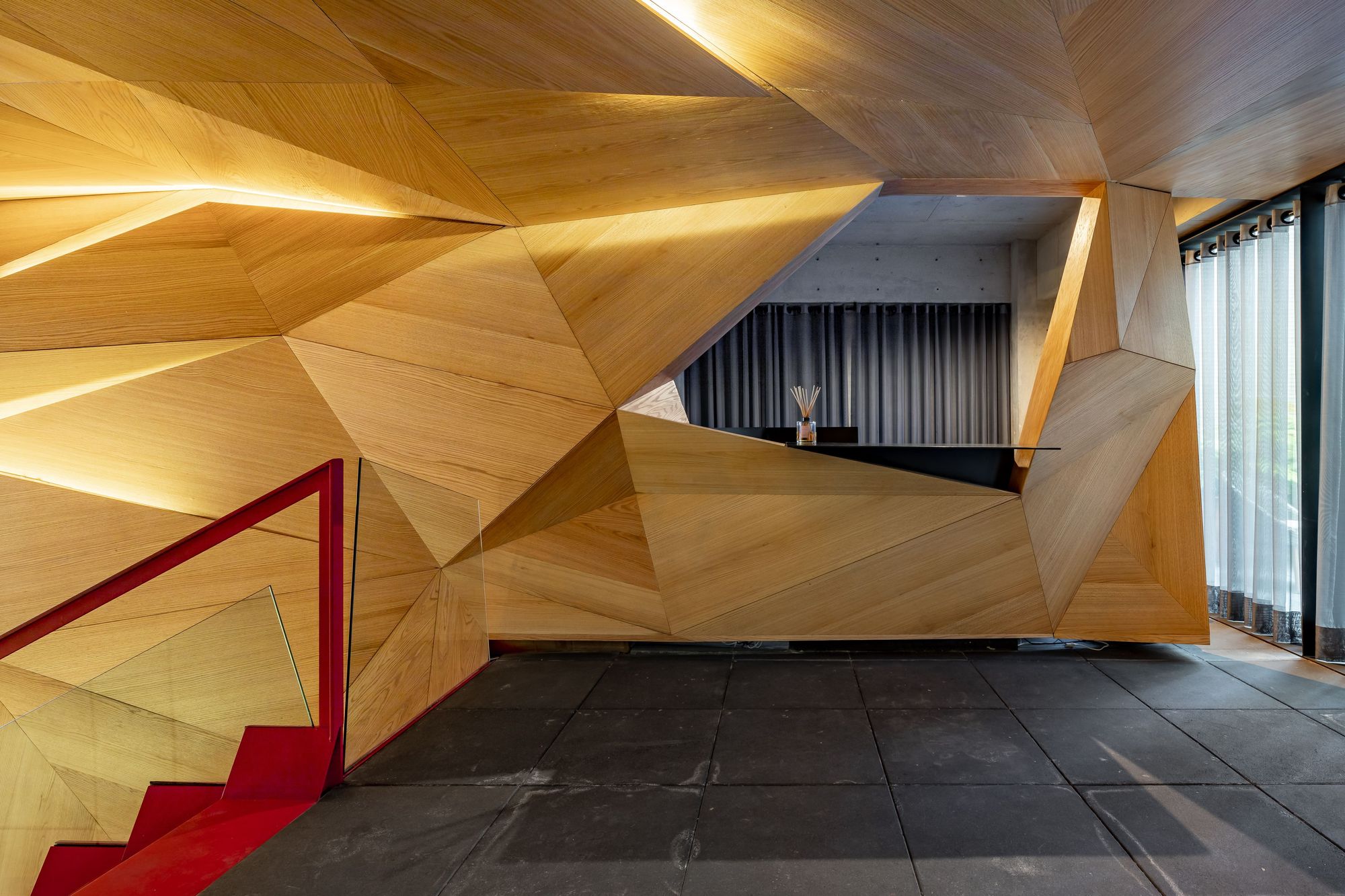 Faceted oak-clad reception desk beside a red-railed stair, lit by warm strip lighting and daylight from curtained glazing