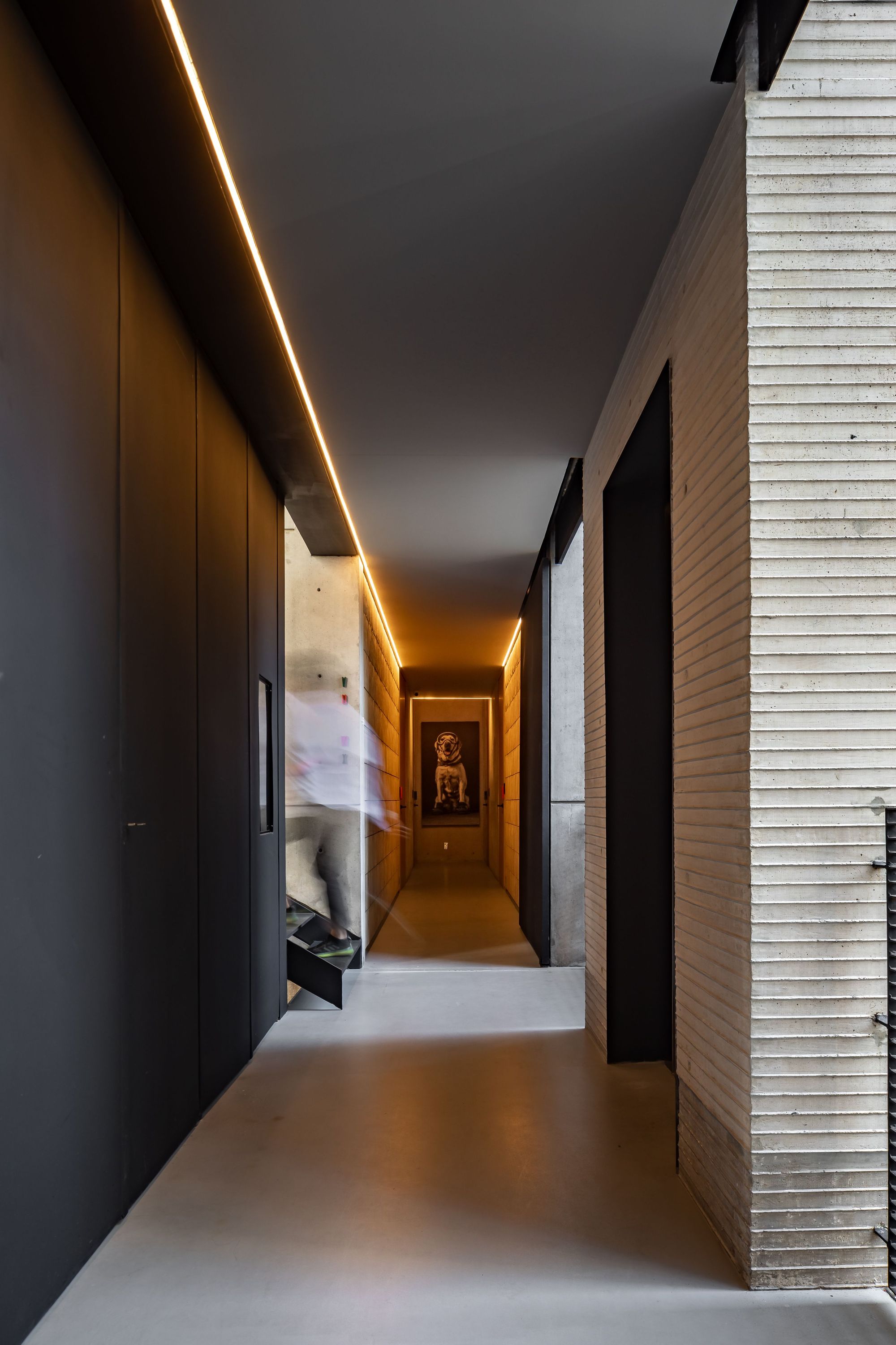 Corridor with a glowing linear ceiling light, dark panelled wall and ribbed concrete column, with a blurred figure passing on the stair
