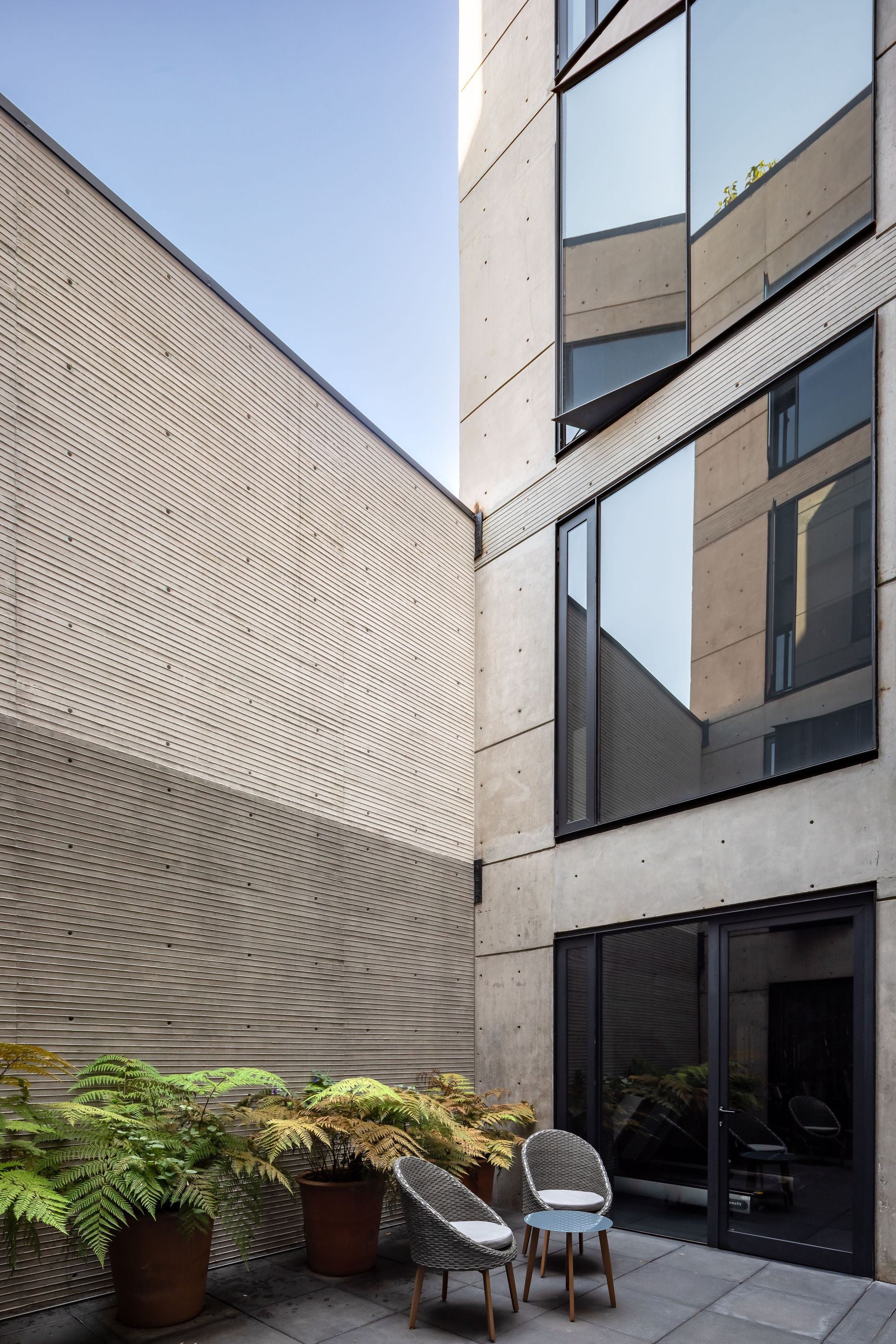 Courtyard terrace with potted ferns and wicker chairs below a ribbed concrete wall and reflective angled windows under clear blue sky