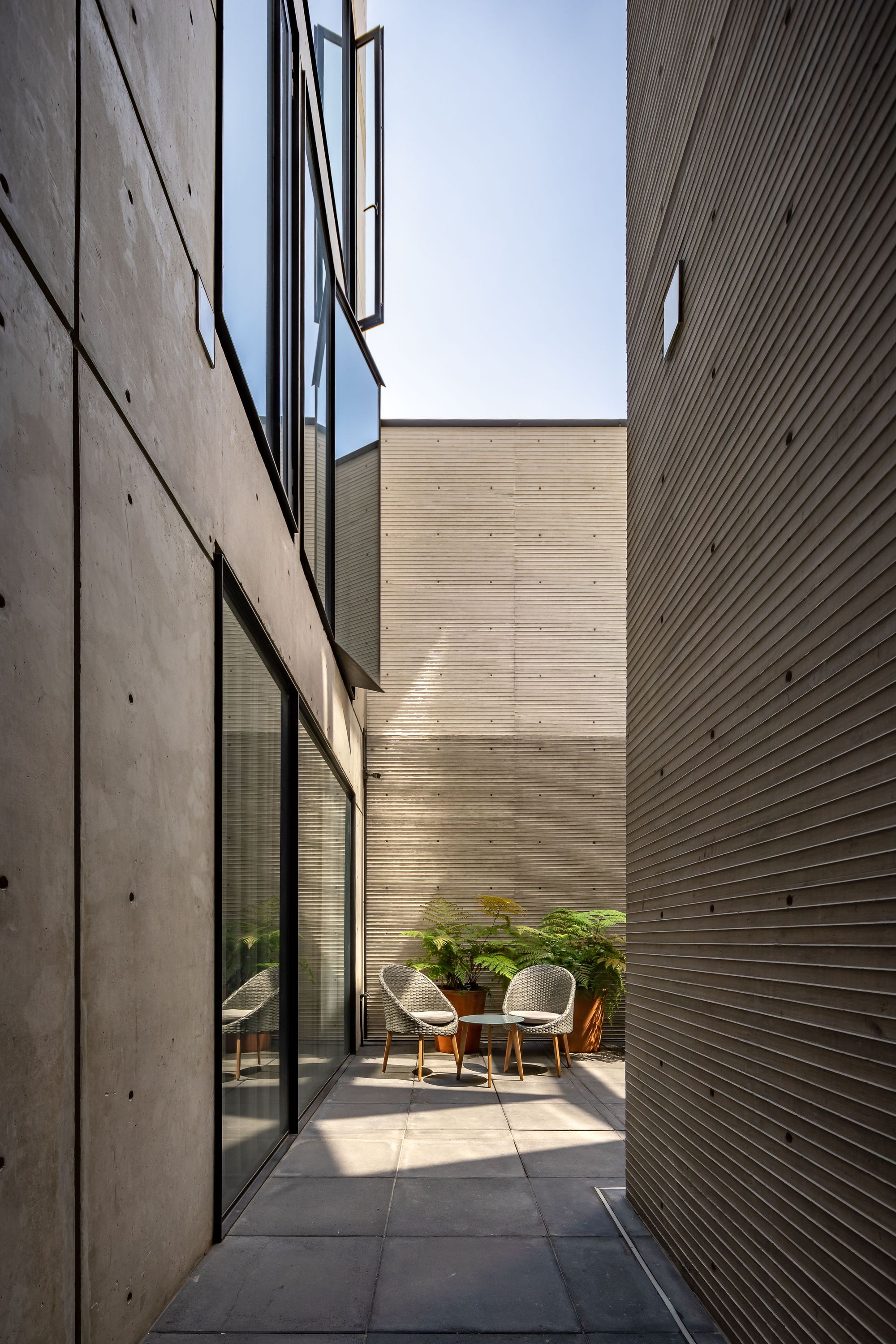 Narrow concrete passage between board-formed walls leading to a paved patio with two woven chairs and potted ferns in sunlight