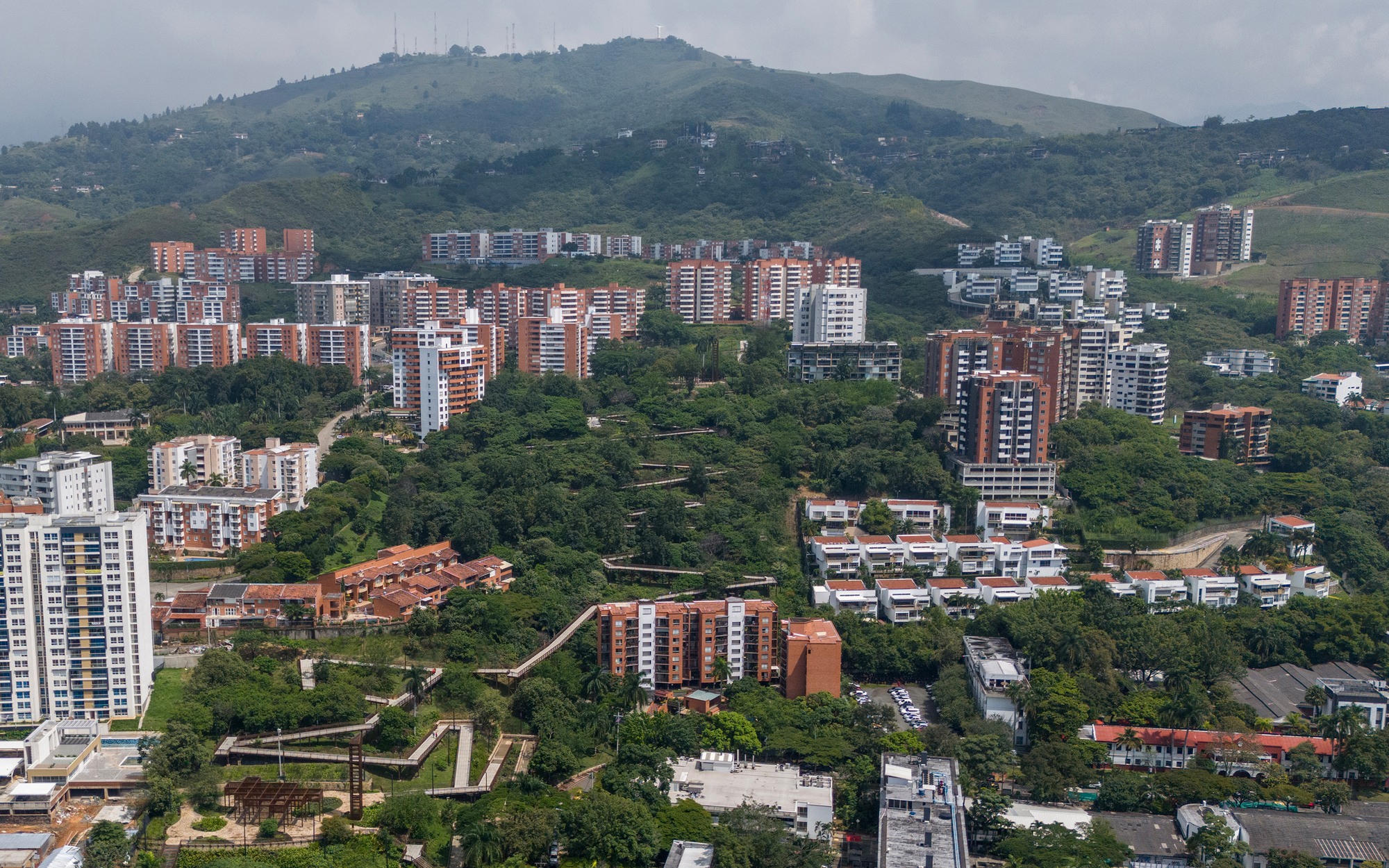 Aerial view of zigzagging elevated walkways cutting through a green hillside park, ringed by brick-colored apartment blocks under overcast sky