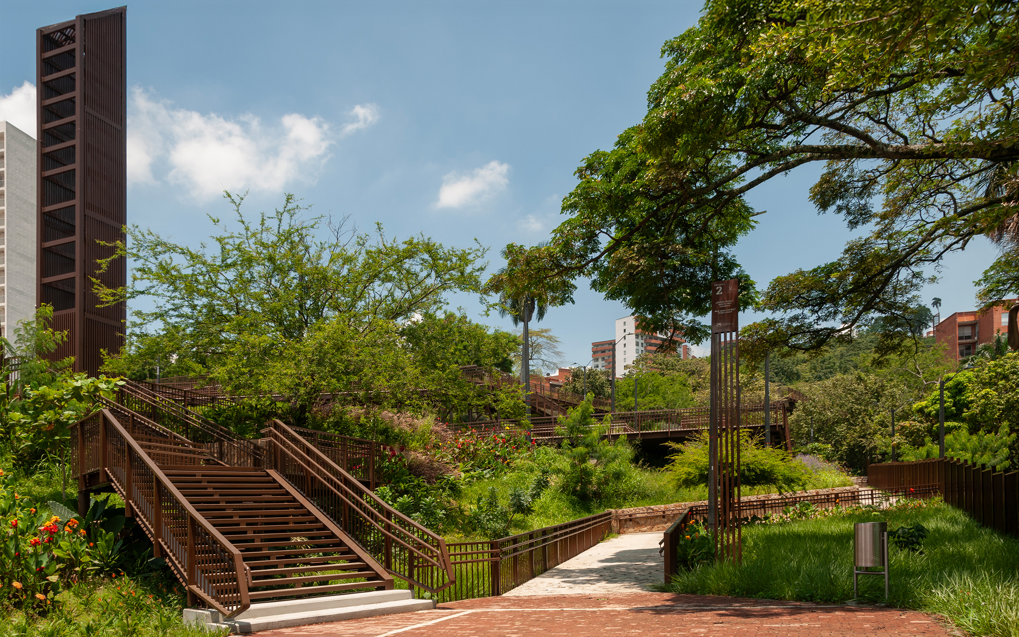 Rust-toned timber staircase and railings rising through planted beds beside a brick path, with a tall slatted tower in blue sky