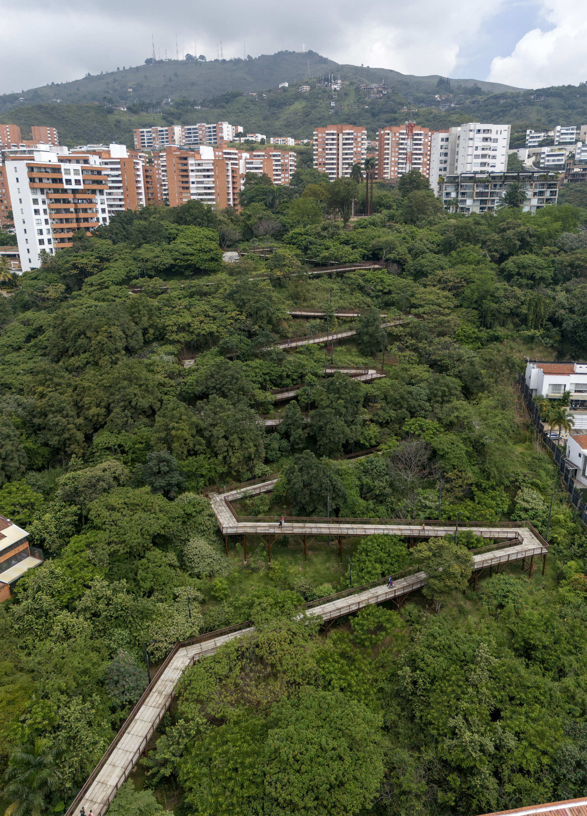 Drone view of switchbacking raised walkways threading through dense tree canopy below apartment towers and a cloudy ridgeline