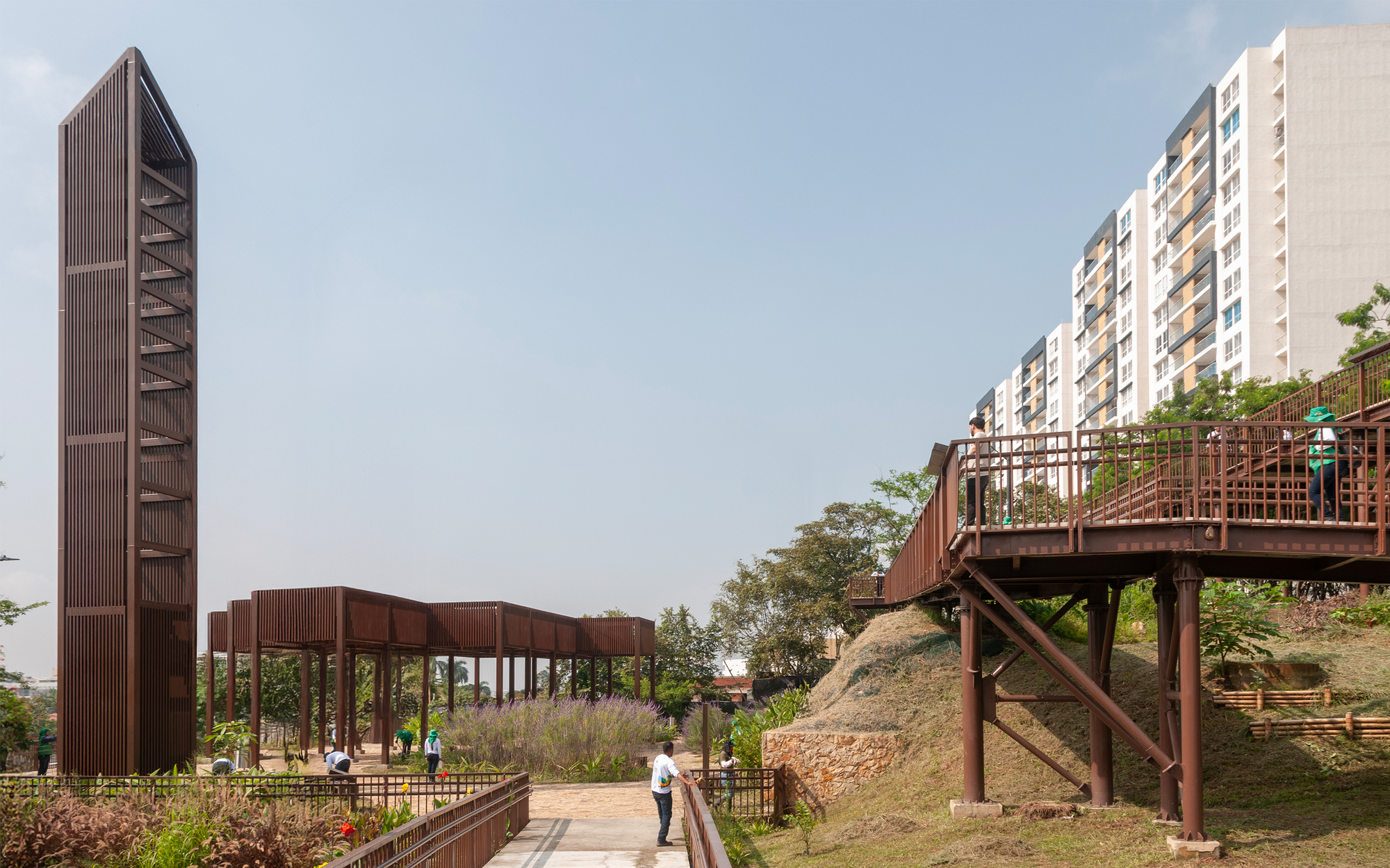 Slatted brown tower beside a steel pergola and a raised steel-framed walkway on an earth embankment, with apartment blocks behind in hazy light