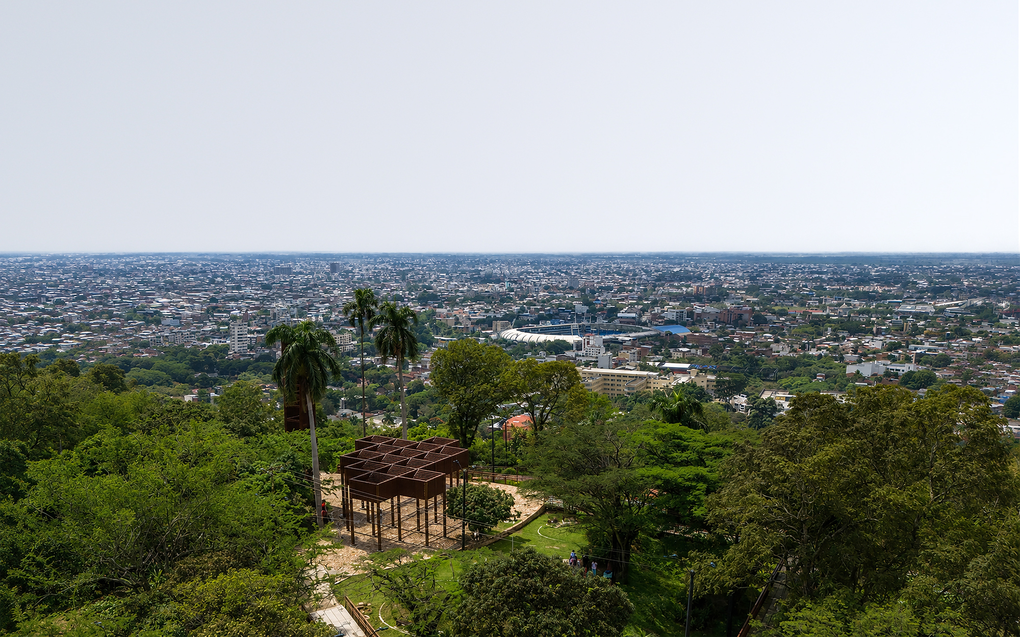 Elevated view of a rusted steel pergola on a wooded hilltop above a sprawling city under hazy sky