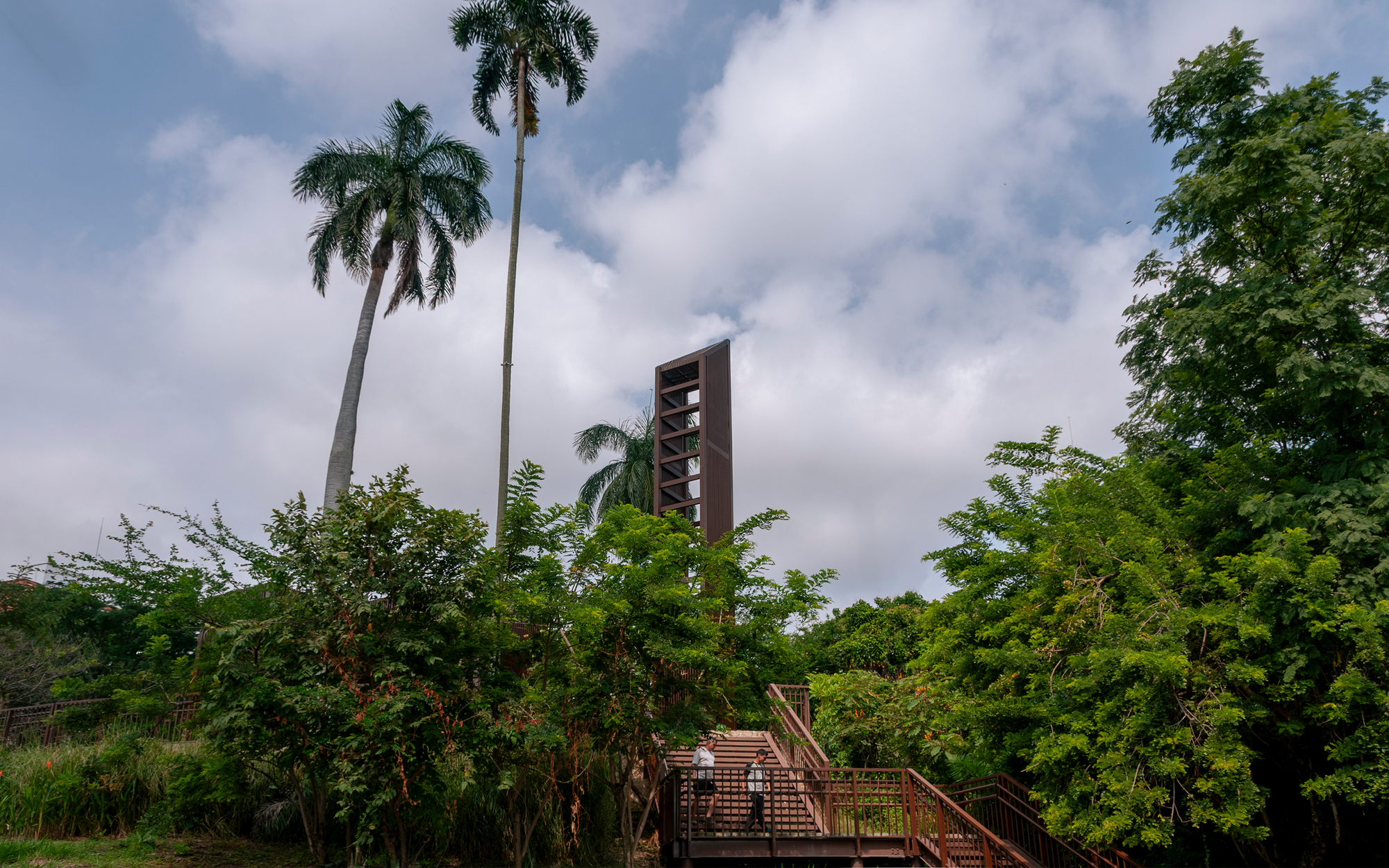 Weathered steel lookout tower rising behind palms and trees, with a stair and two visitors below under cloudy sky