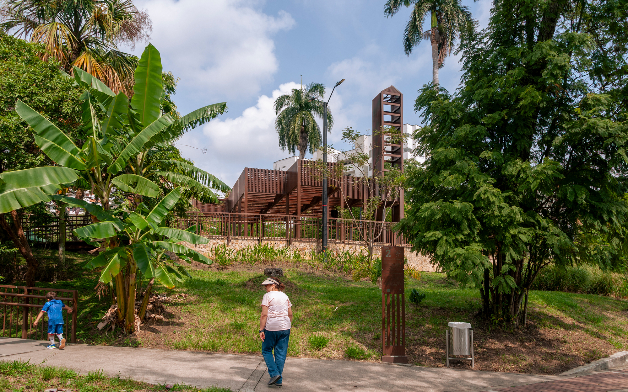 Slatted steel pavilion and tower behind a grassy slope, banana plants and a woman walking the footpath in front