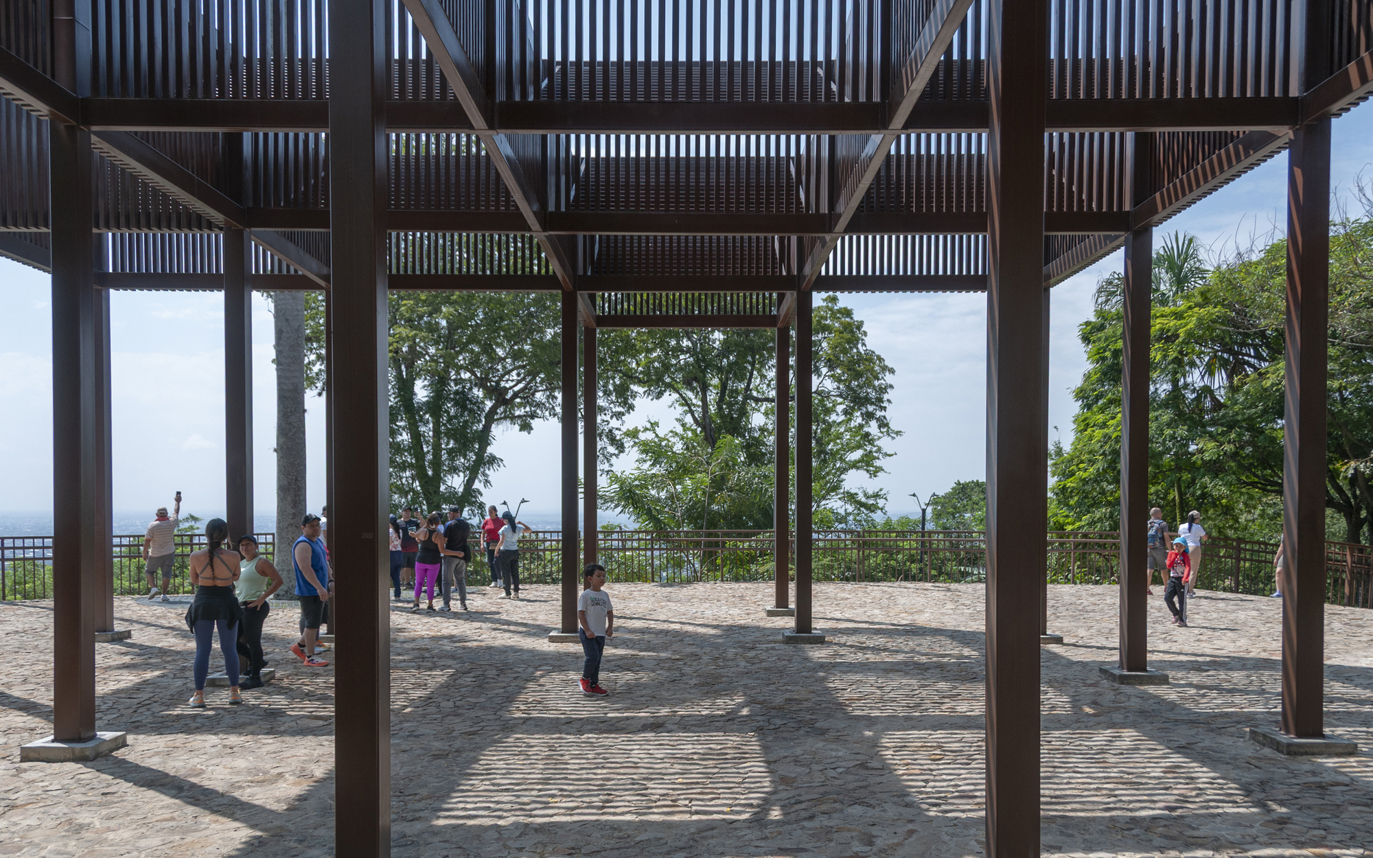 Underside of a timber-framed pavilion with slatted roof above a cobbled plaza, visitors standing in dappled sunlight