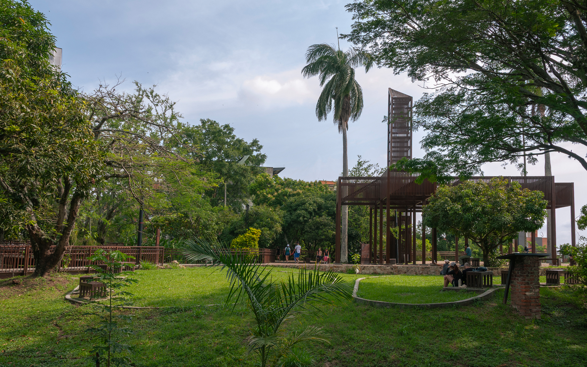 Rust-colored timber pavilion with a tall lattice tower rising behind a palm, across a lawn with curved edging and brick pillar