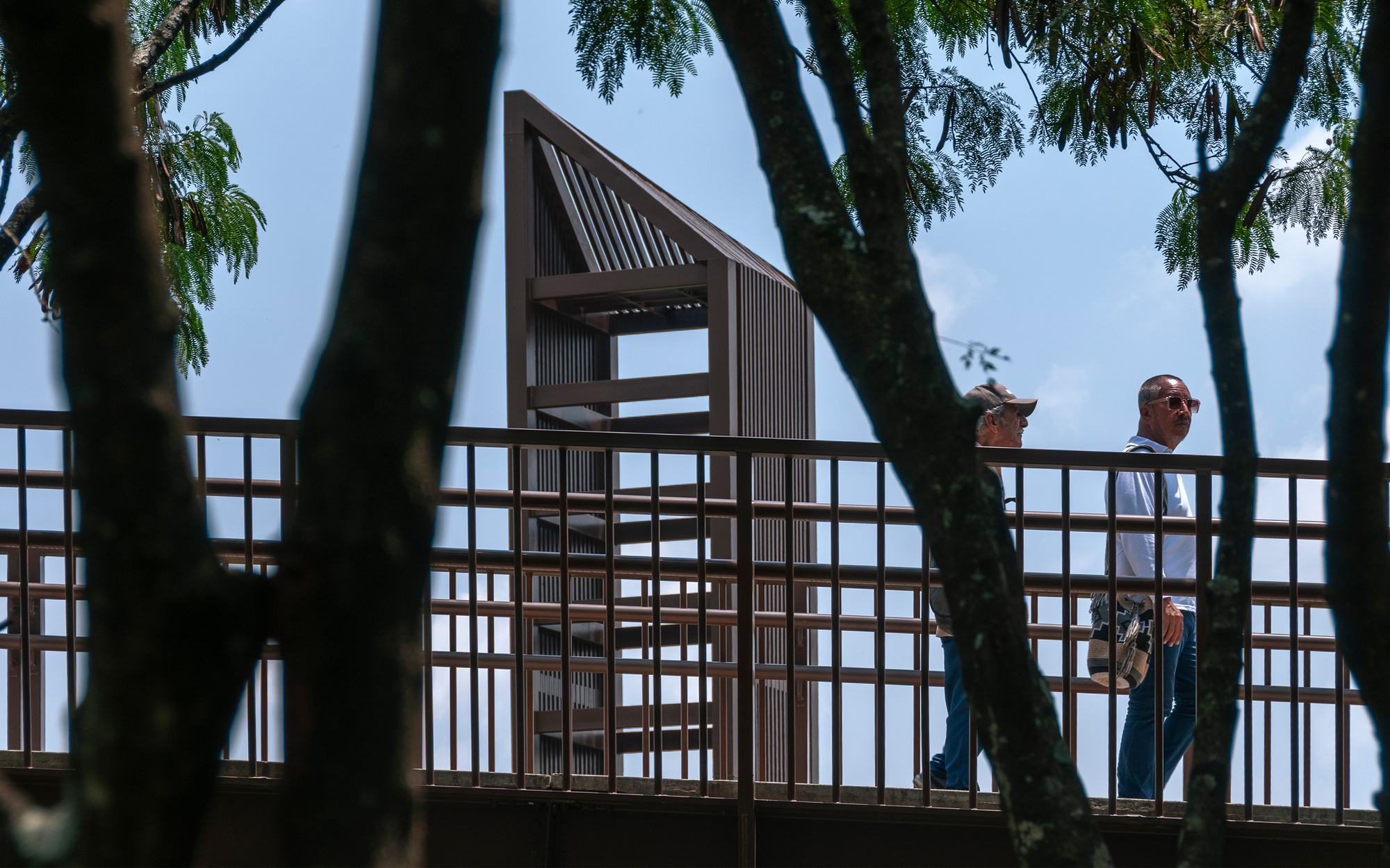 Two walkers cross a steel-railed elevated path beneath tree trunks, with a slatted timber tower rising against a blue sky