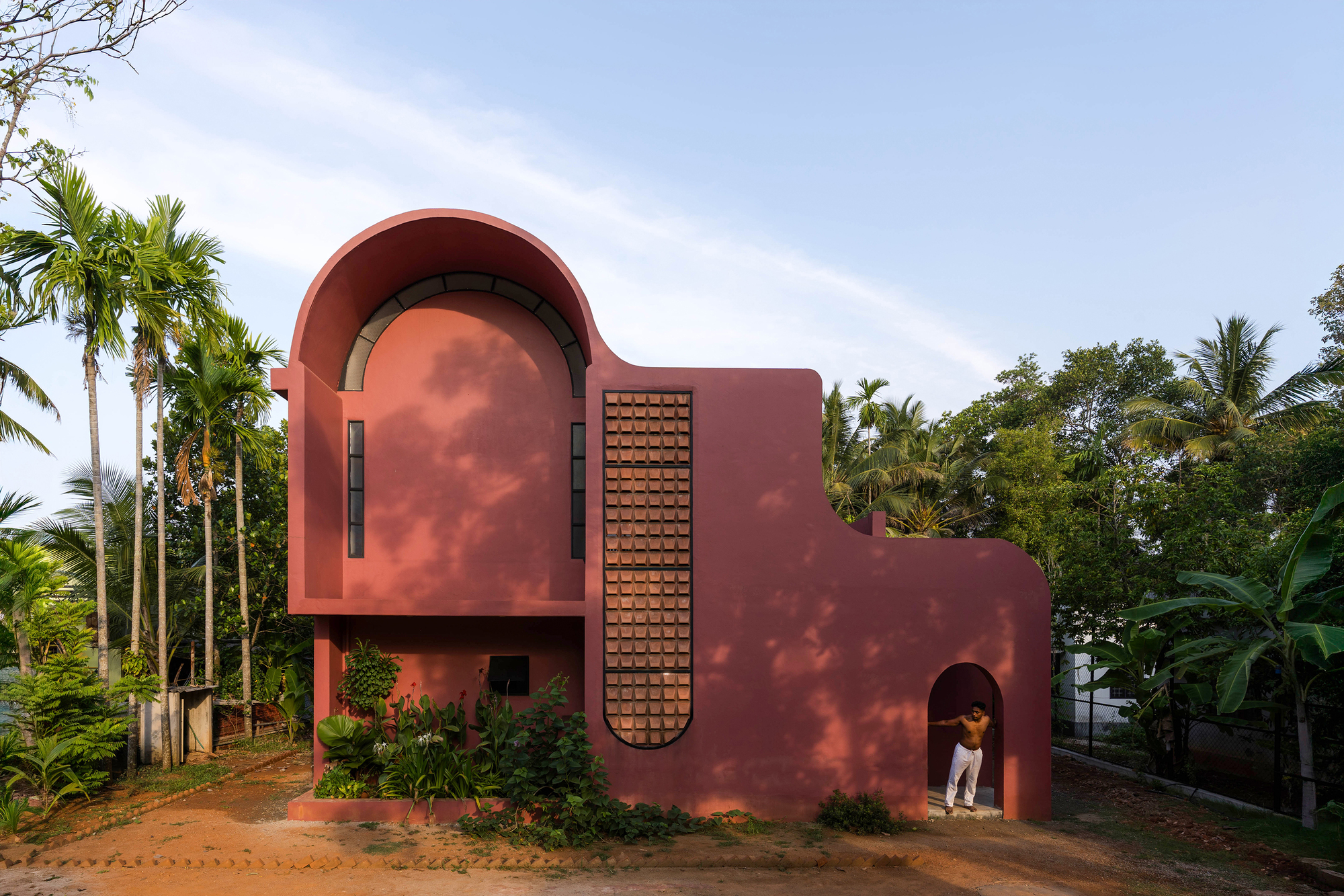 Red rendered facade with an arched upper volume and tall perforated brick screen, a man standing in the arched doorway beside palms