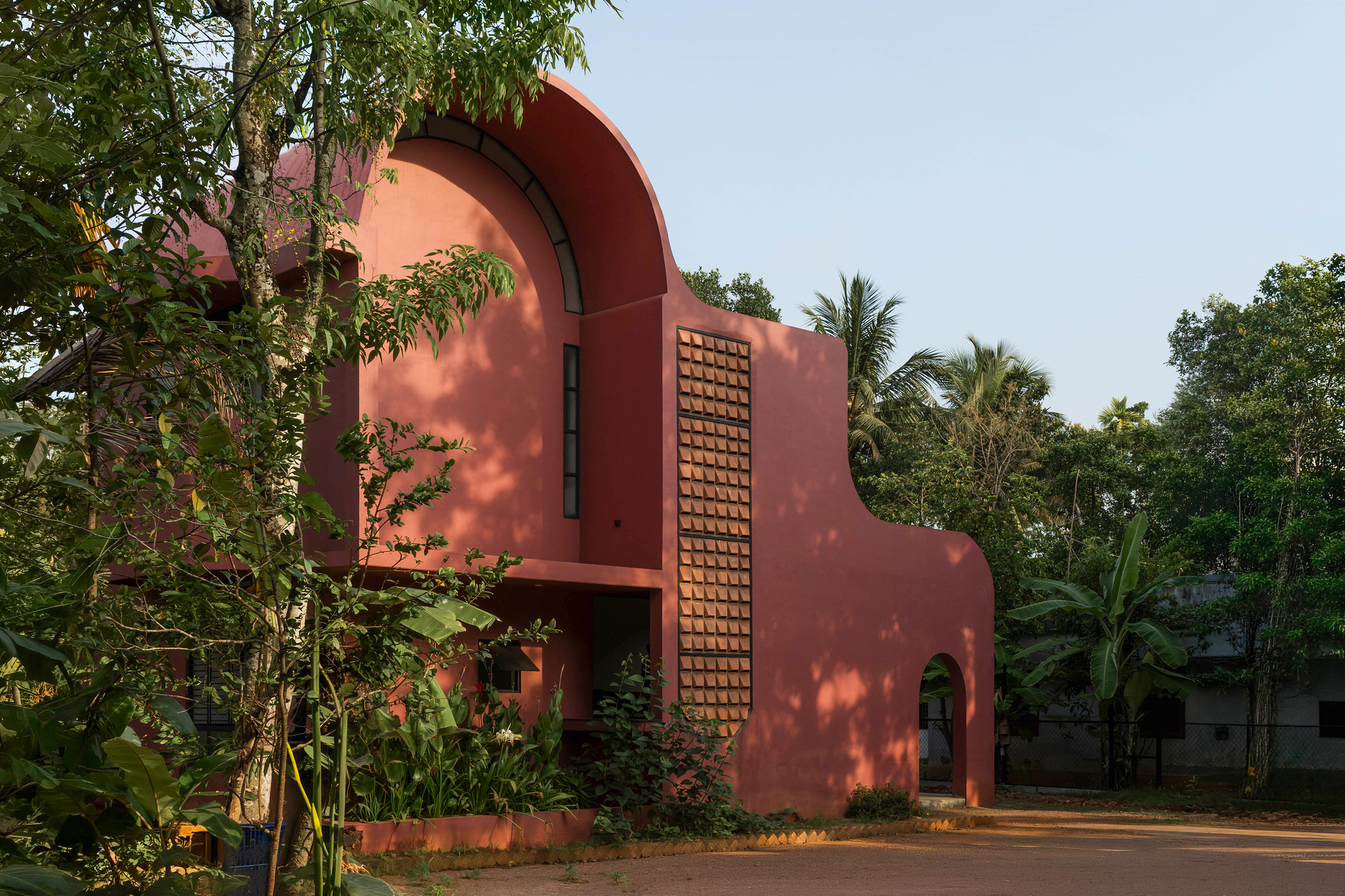 Side view of the red curved facade and brick screen, dappled with leaf shadows from foreground trees in afternoon light