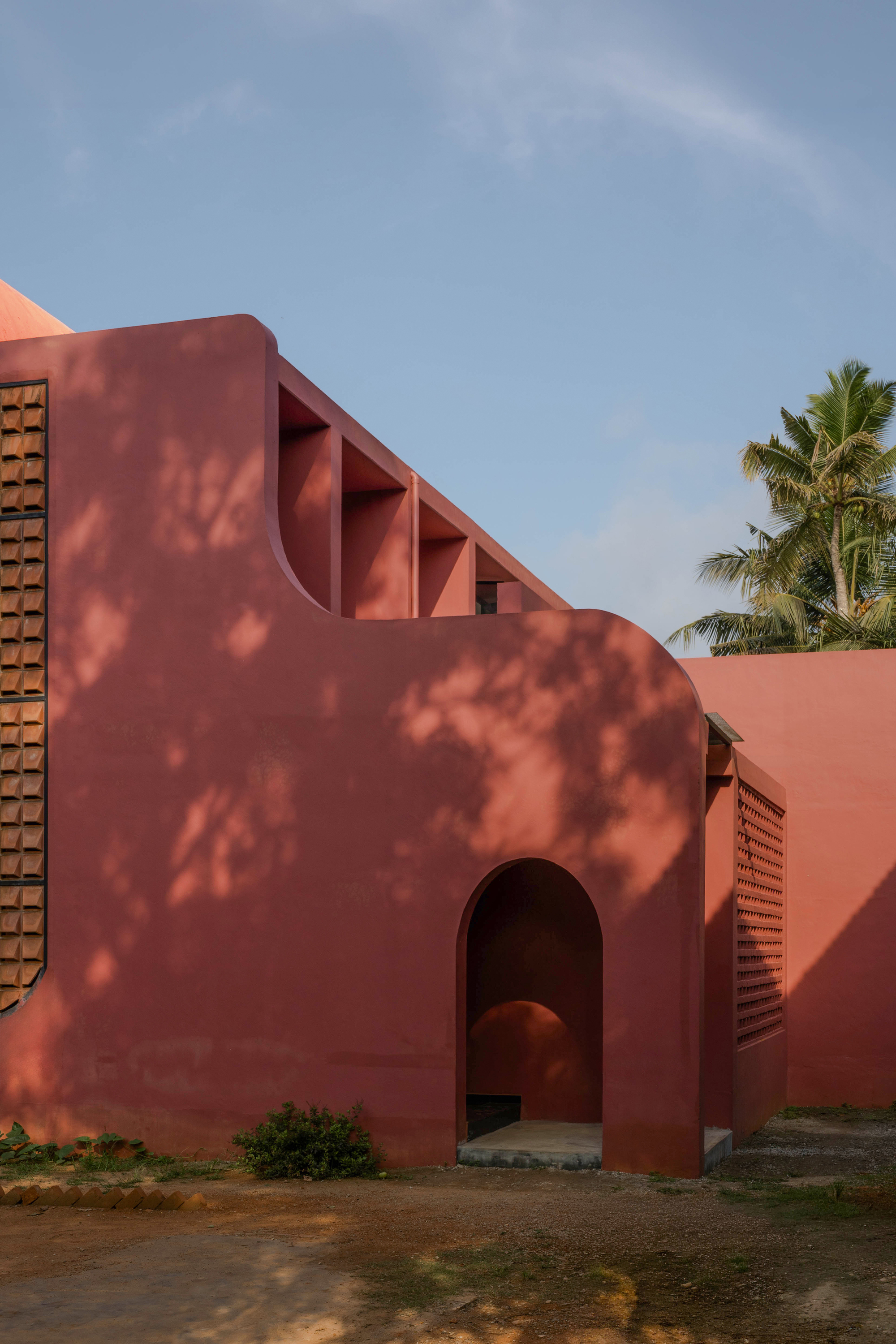 Rounded red wall corner with a small arched entrance and stepped openings above, shadows of palm leaves falling across the plaster