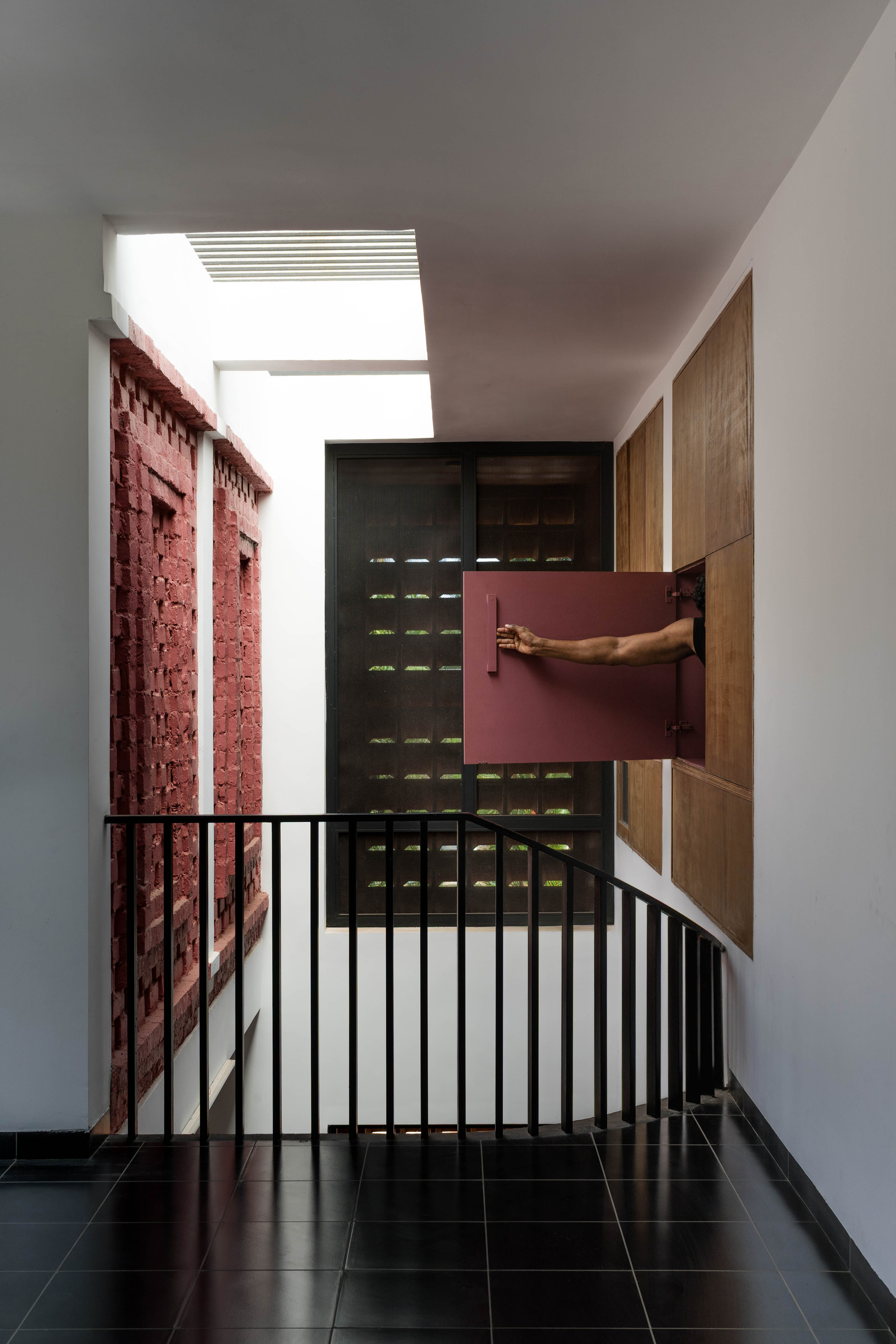 Stairwell landing with black balustrade, red brick lattice wall and a hand pushing open a red wooden shutter beneath a skylight