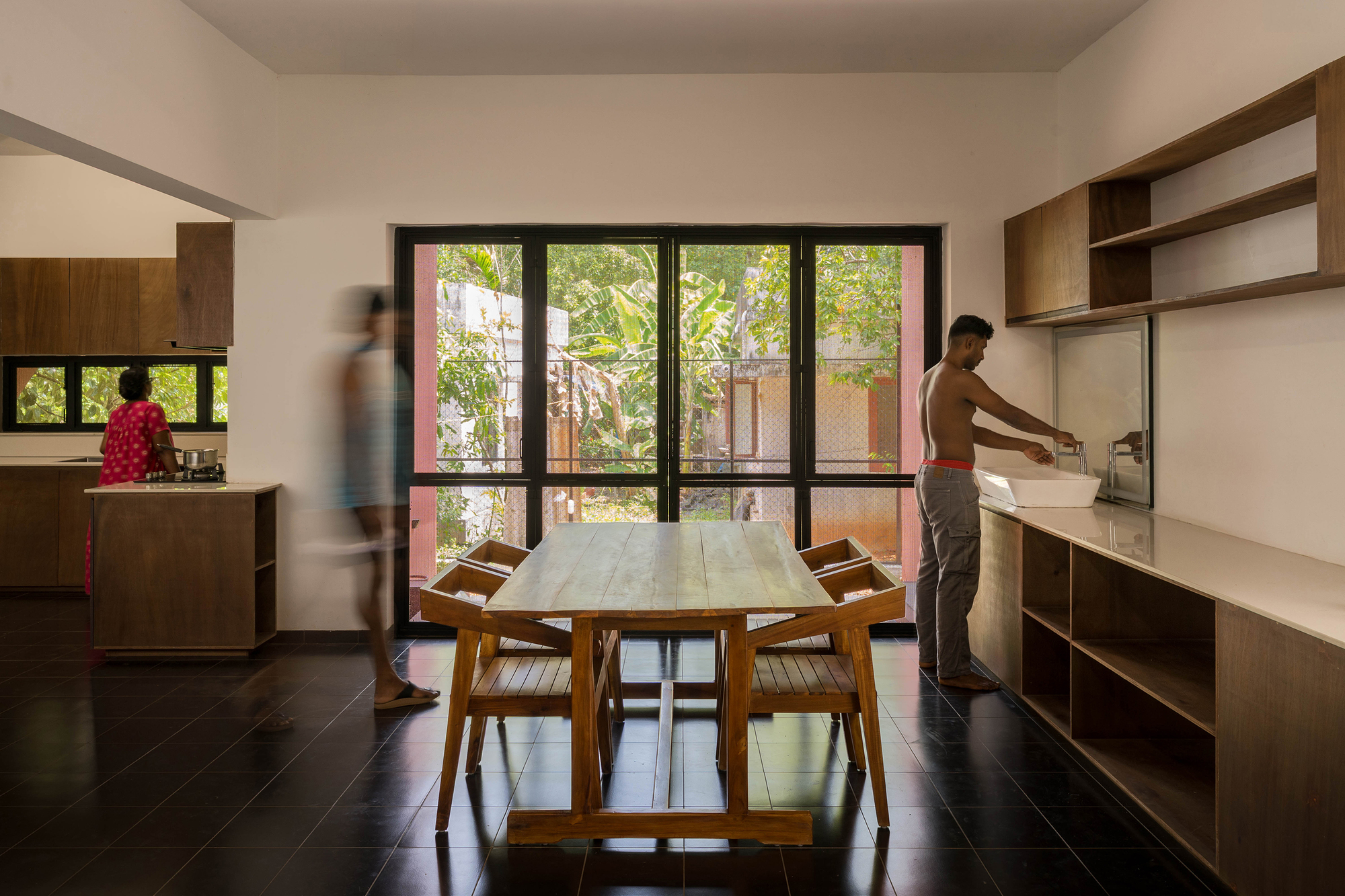 Dining area with a timber table and black-framed glass doors, a blurred figure walking past and a man washing at the counter