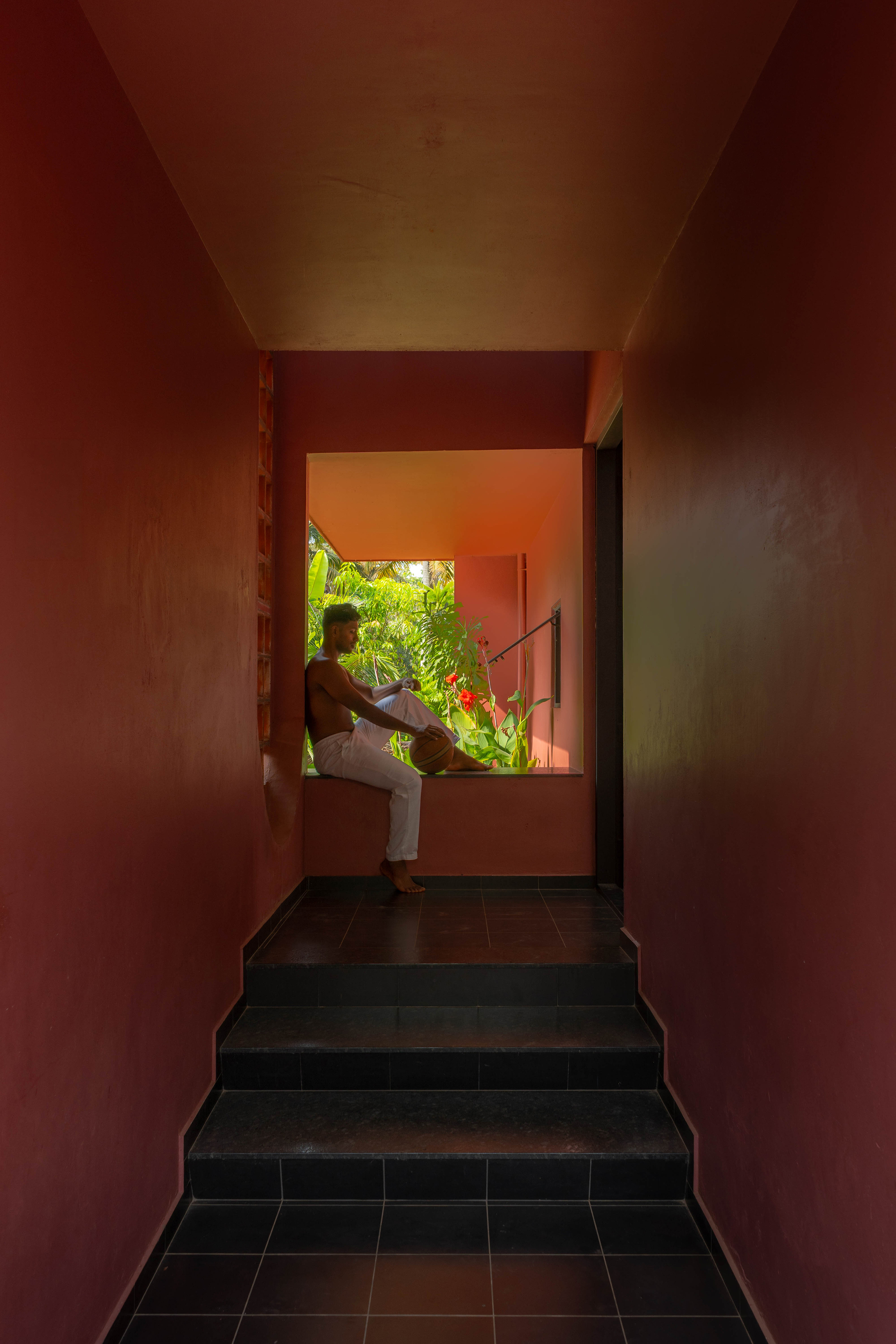 Narrow maroon corridor with black stone steps leading to a window seat where a man sits against tropical greenery