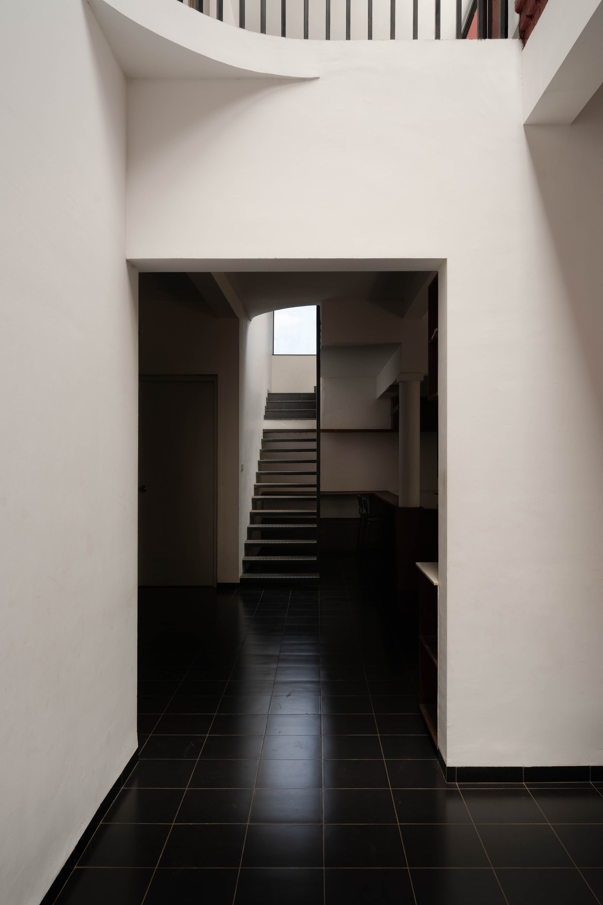 White plastered opening framing a dark tiled floor and a steel staircase rising toward a skylight in low light
