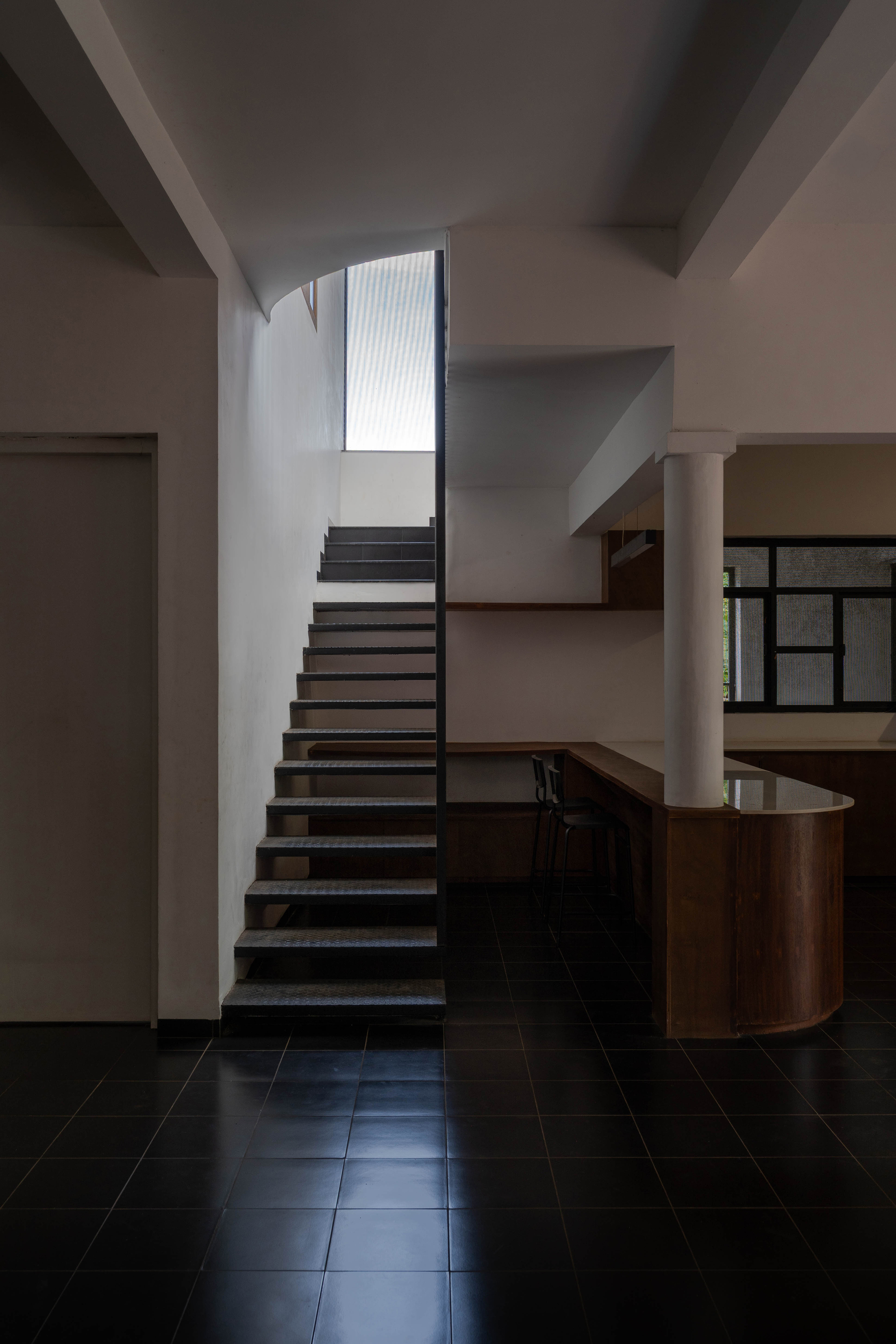 Open-tread steel staircase beside a round white column and a curved timber counter on a black tiled floor