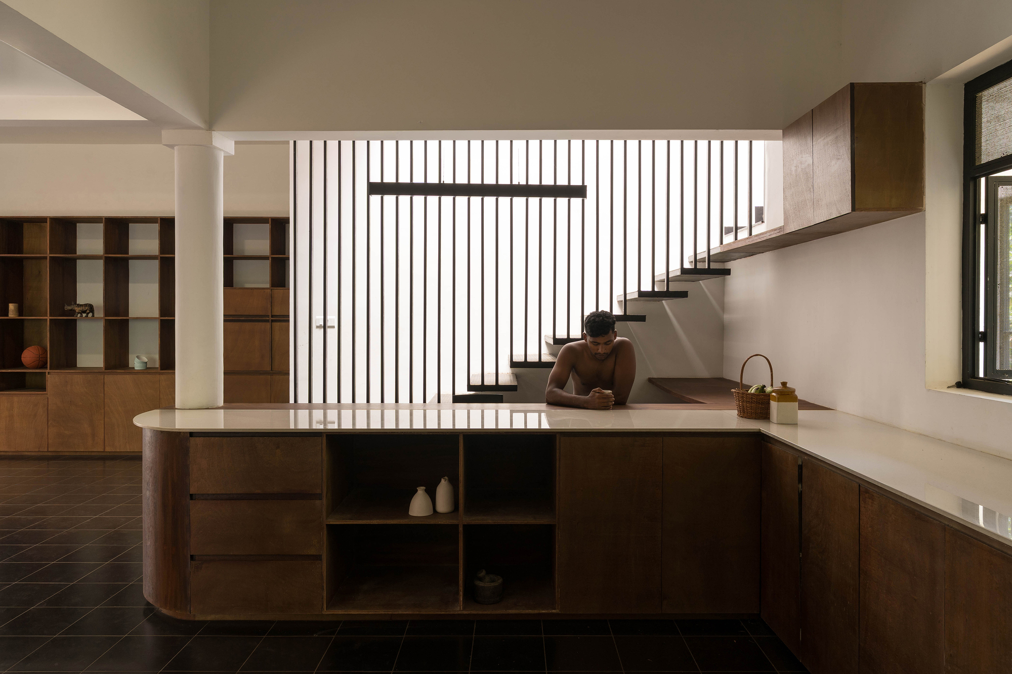 Curved timber kitchen counter with open shelves, a man leaning on it before a slatted stair screen