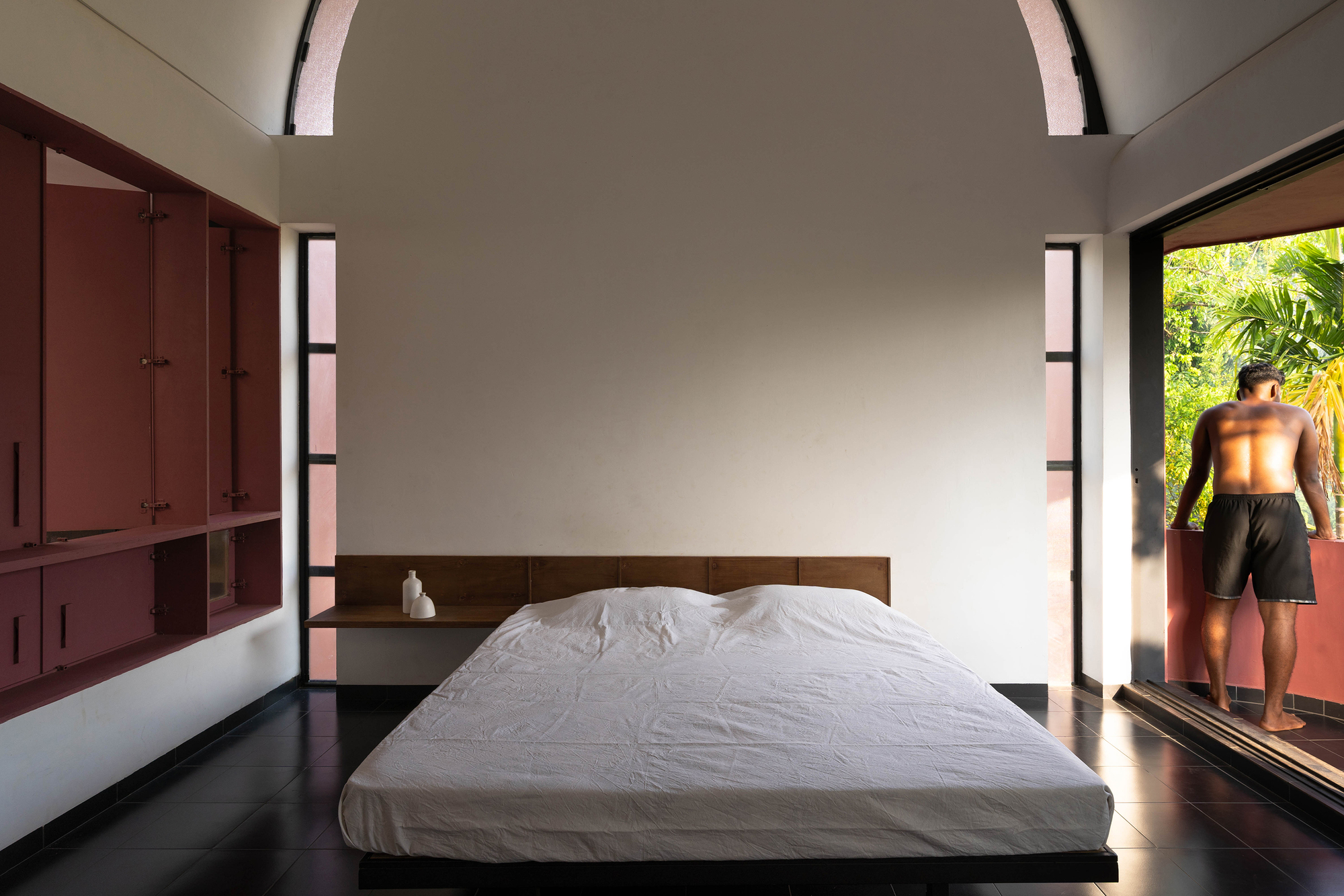 Bedroom with a low white bed, maroon built-in cabinets and a barefoot man on the balcony among palms