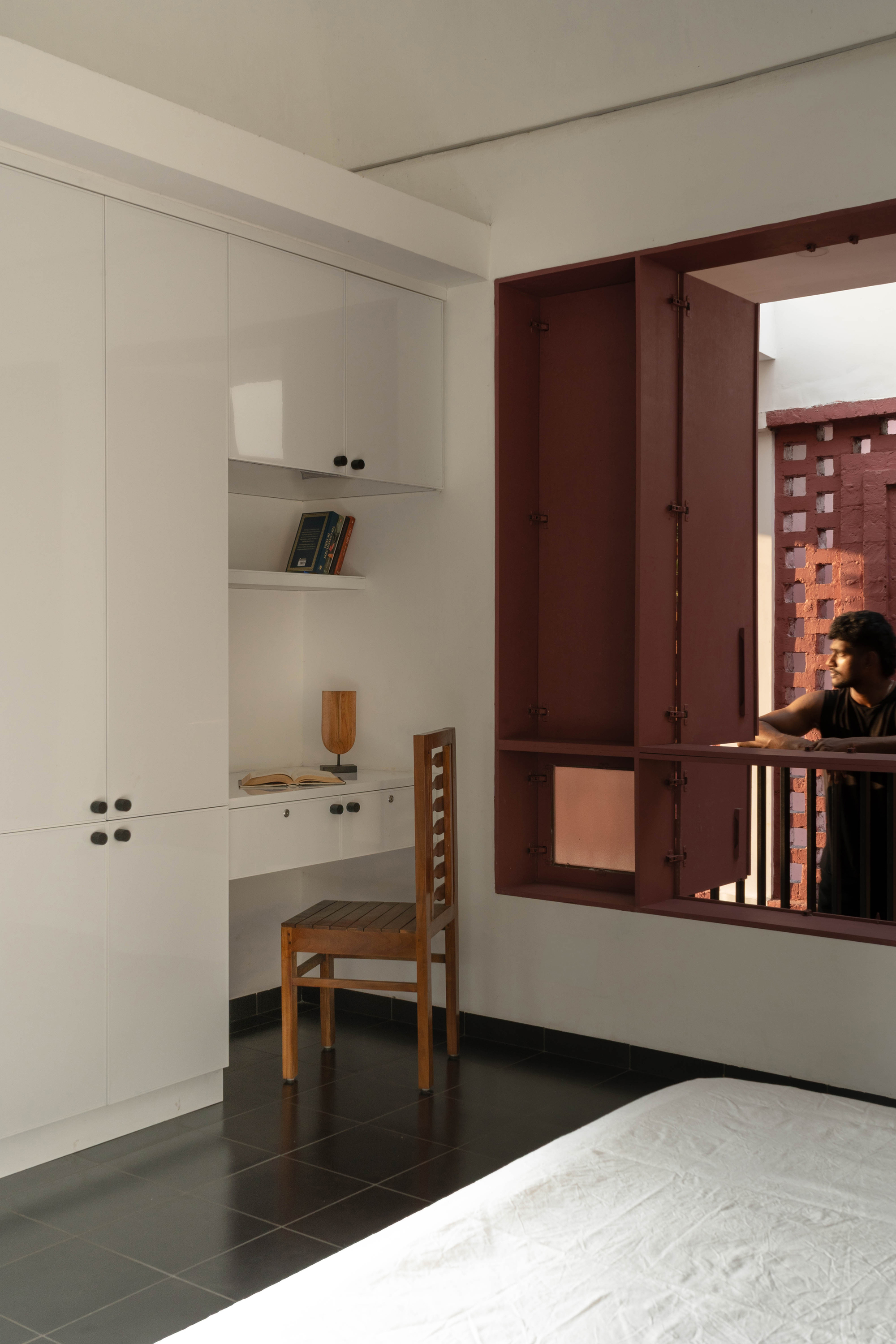 Corner of a bedroom with white cabinets, a built-in desk, a wooden chair and a maroon shuttered window with a man looking out