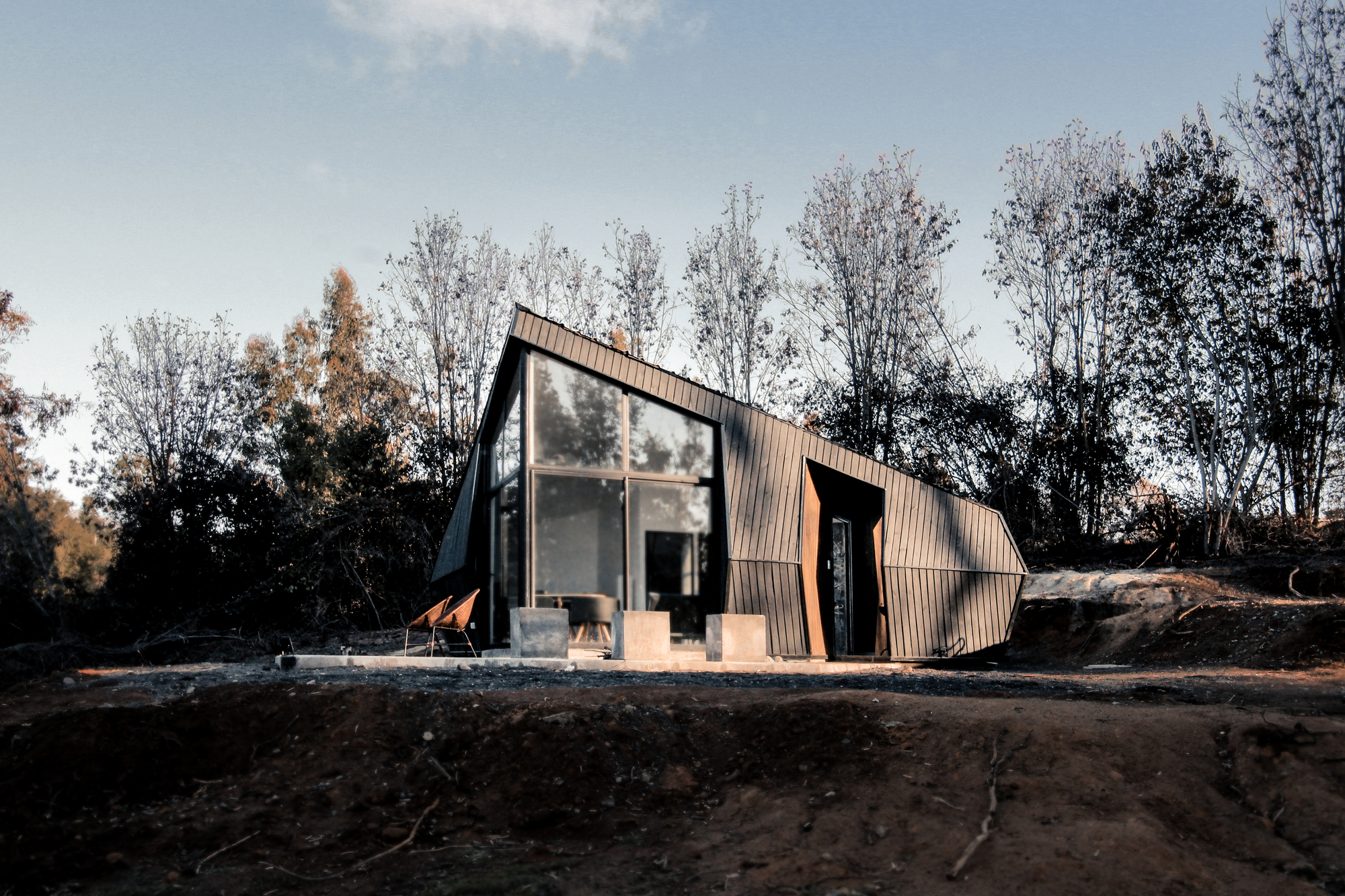 Dark timber-clad cabin with a glazed gable end and angled entry recess, set on bare red earth among bare trees at dusk