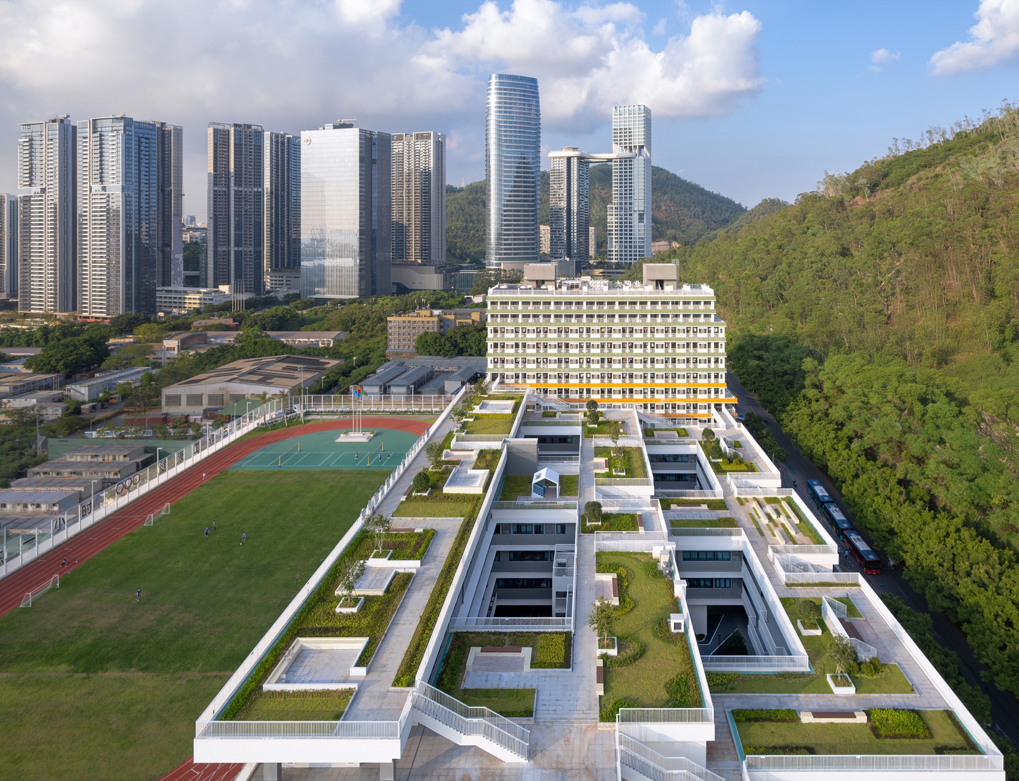 Aerial view of the stepped green roof terraces with courtyard voids, a running track and a forested slope beside high-rises