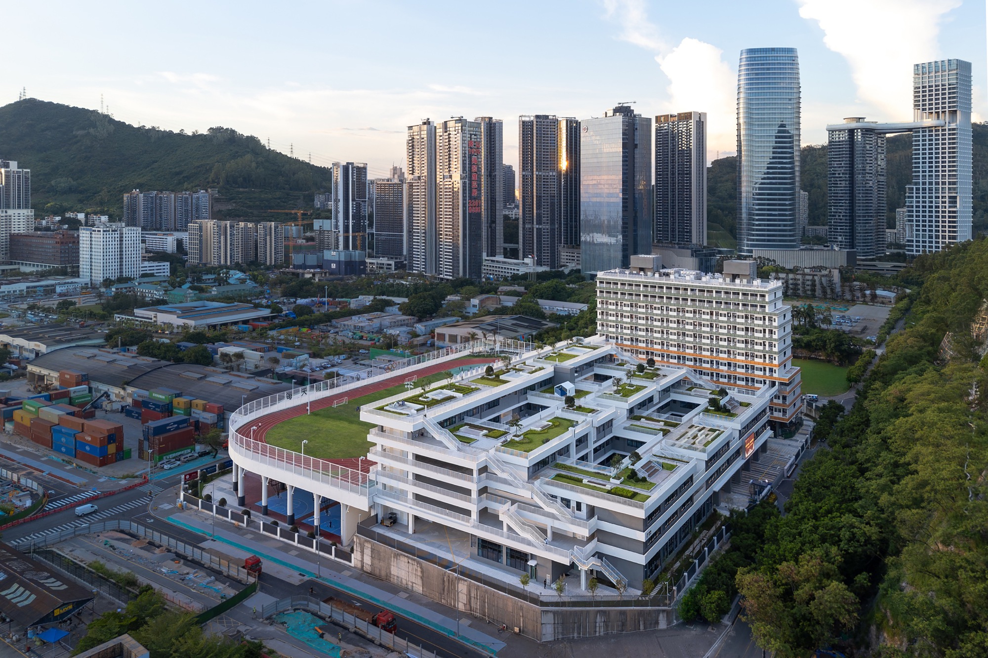Aerial view at dusk of the terraced school block with rooftop track, planted decks and a slender tower behind