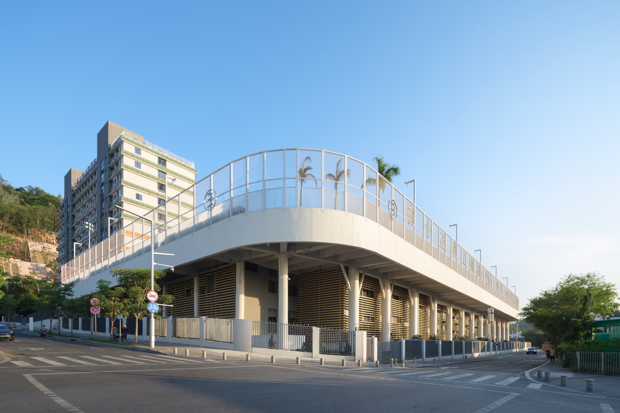 Street corner view of the curved white podium edge on slender columns above timber-slatted walls under a clear blue sky
