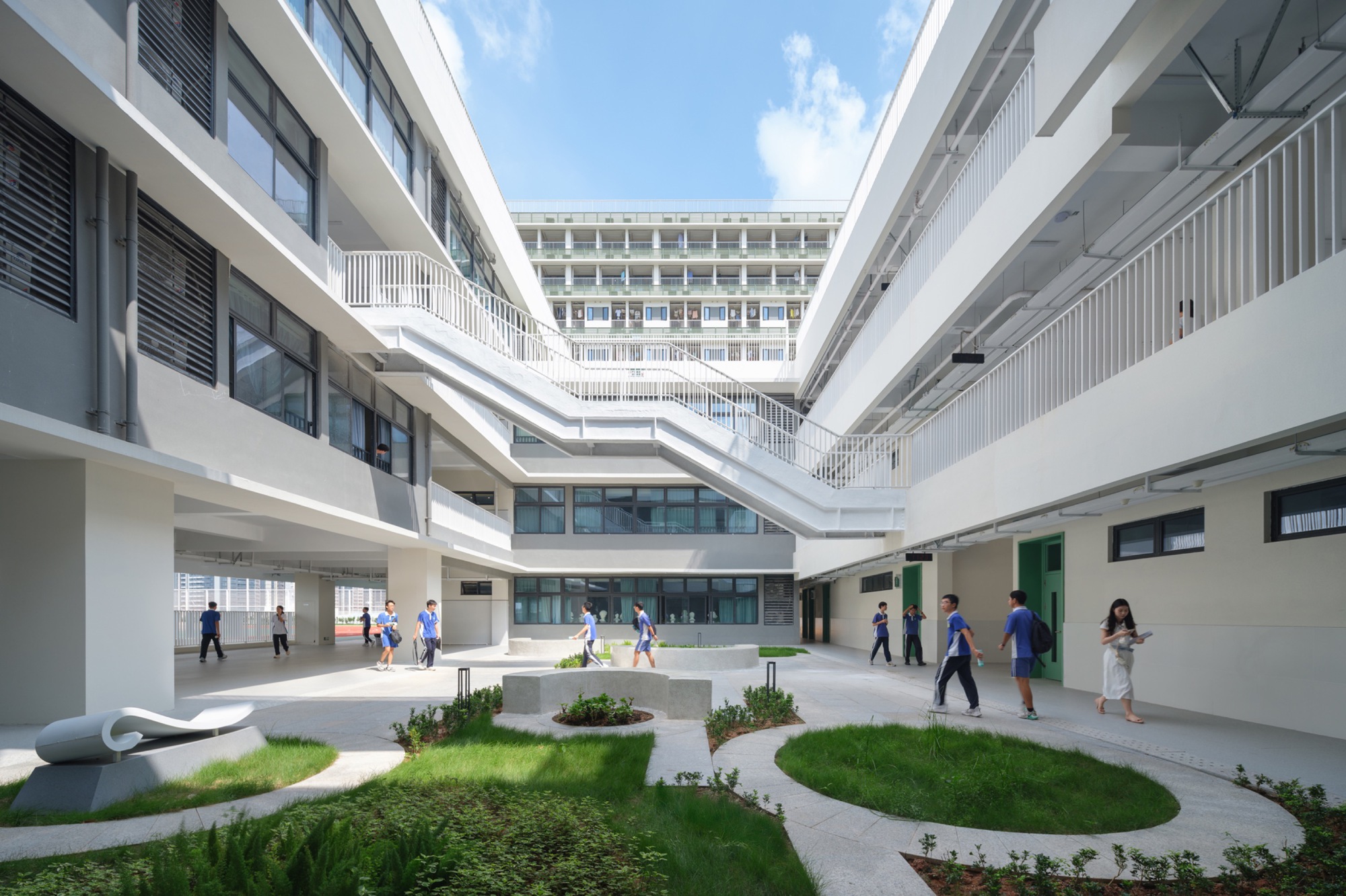 Inner courtyard with white open stairs, circular planted lawns and students walking across the paved floor beneath a partly cloudy sky