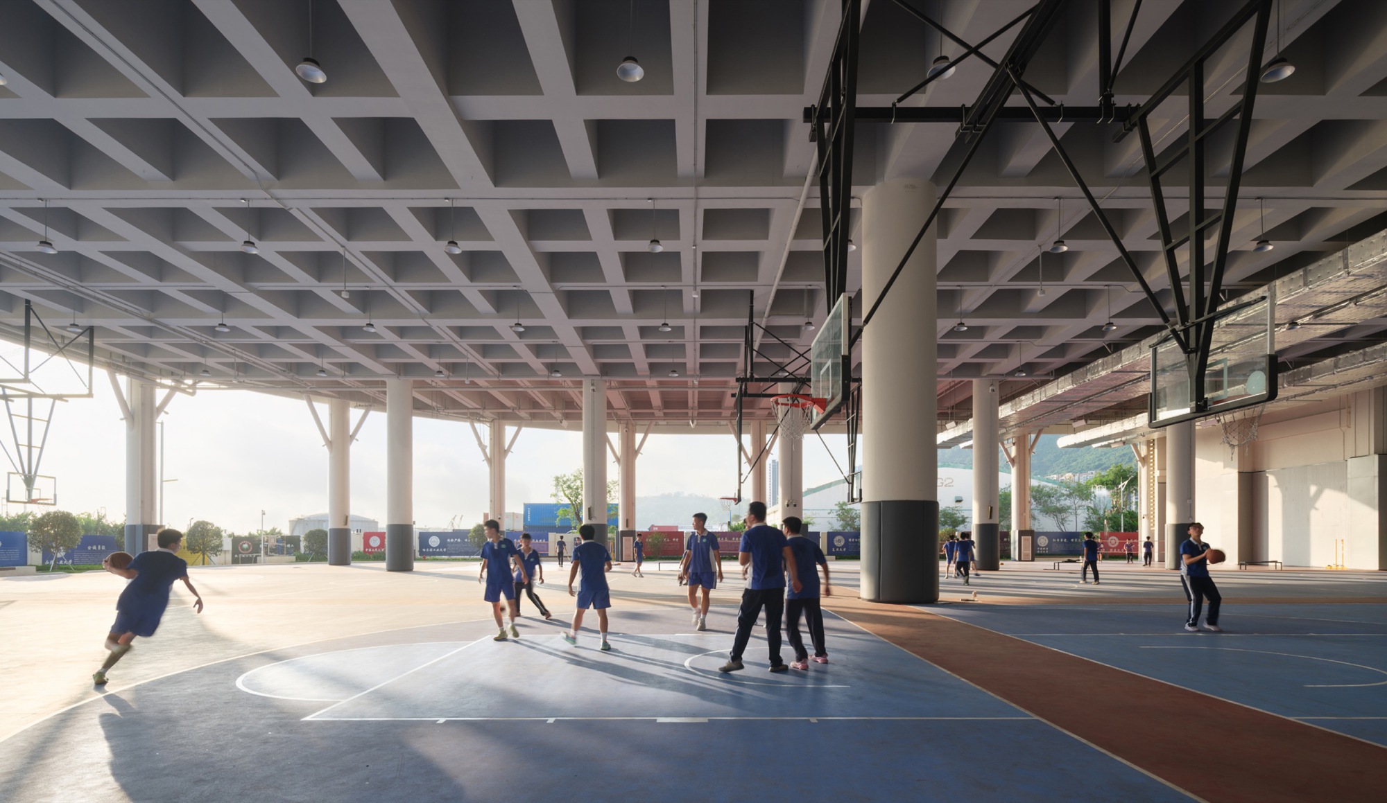 Covered ground-level basketball court under a coffered concrete ceiling with round columns and students playing in late light